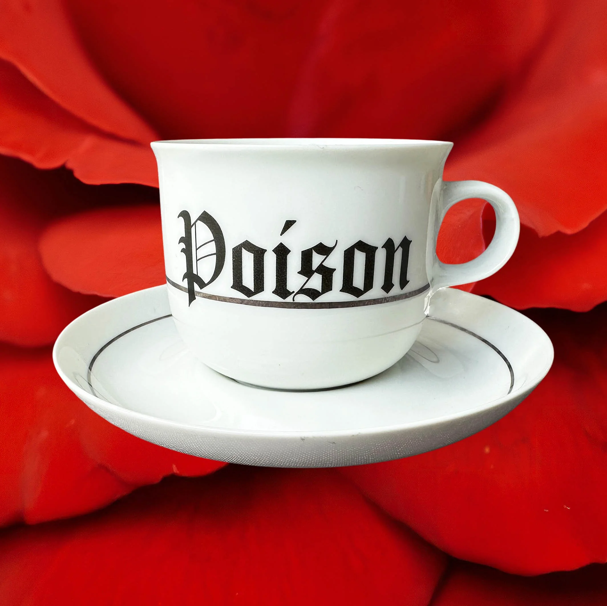 A vintage white tea cup and saucer with a silver band, with the words “Poison” in black gothic style lettering