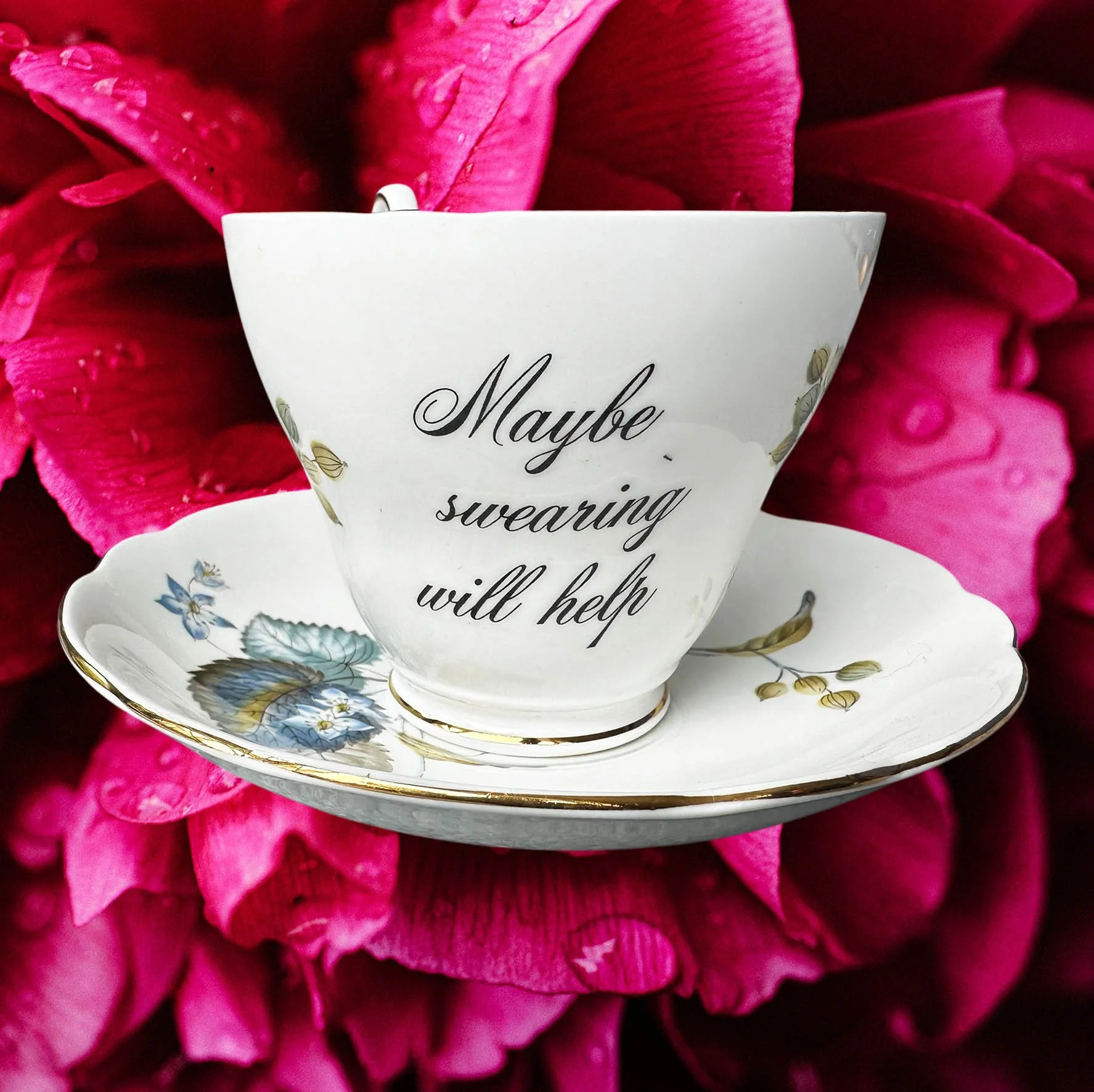 A vintage tea cup with a blue floral design and the words "maybe swearing will help" in black writing