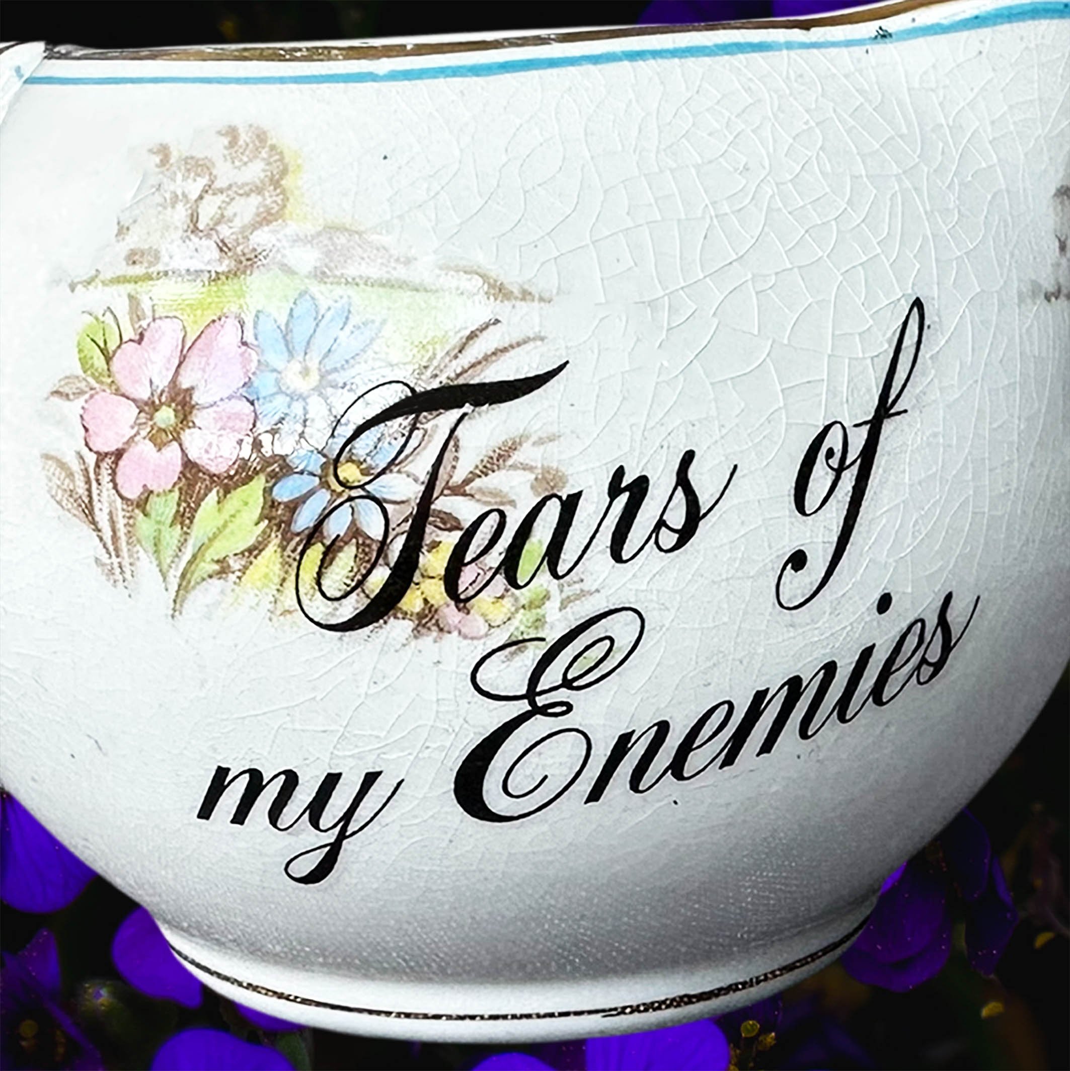 A cream coloured vintage milk jug with a floral design, the words “tears of my enemies” in black cursive writing