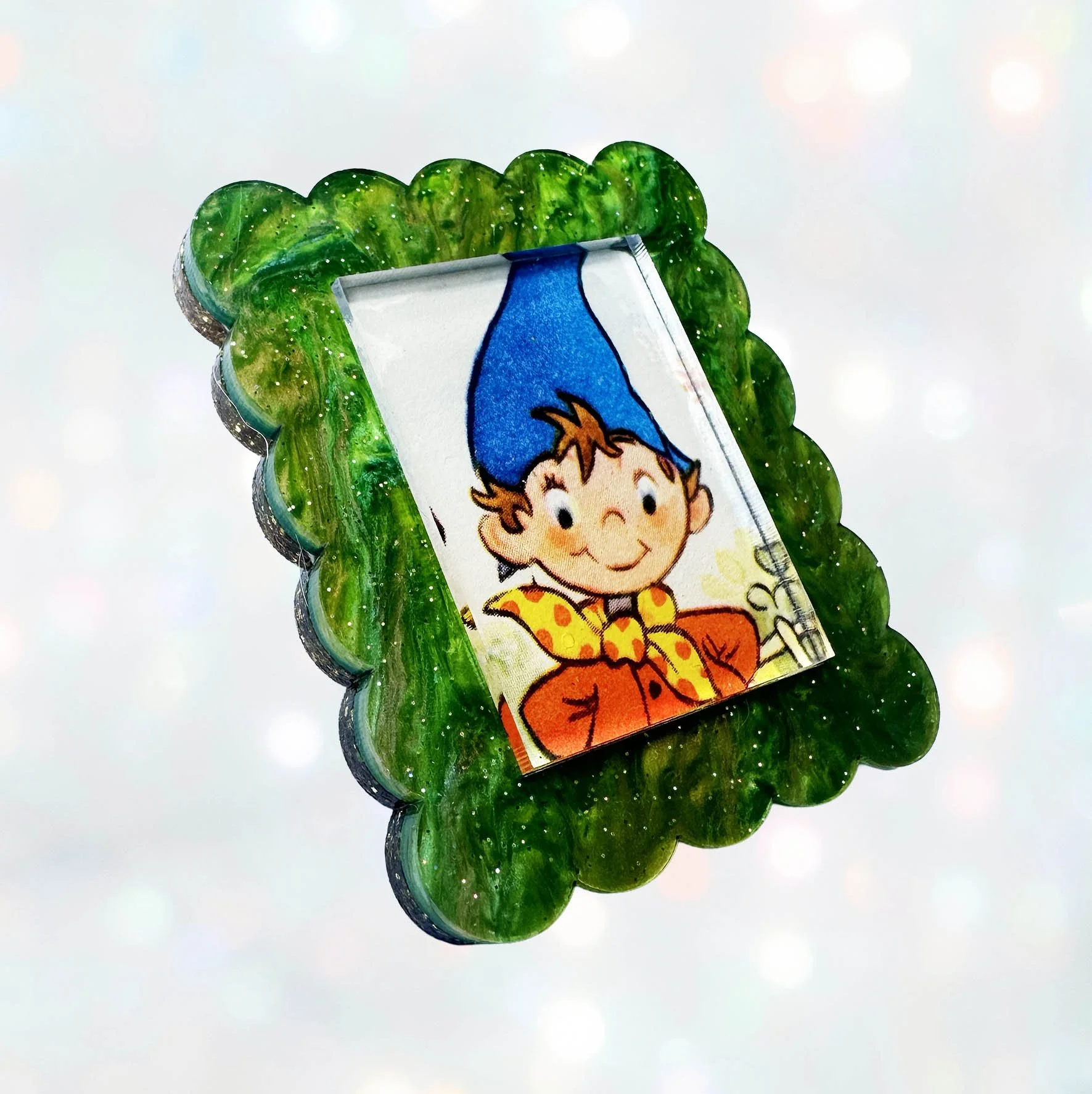 A green glitter acrylic brooch featuring a picture of Noddy