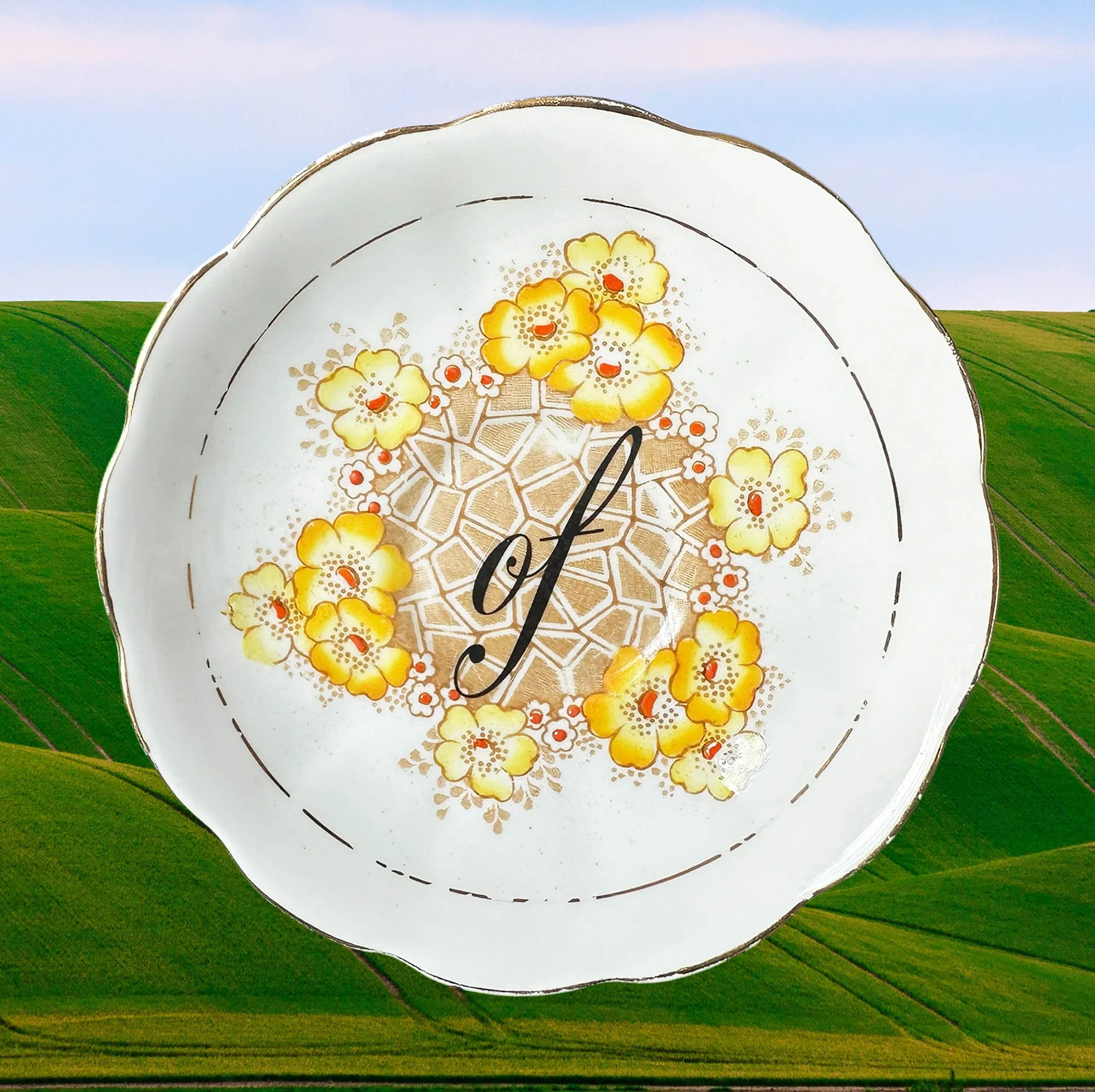 A vintage saucer with a yellow floral design with the words “of” in black cursive writing