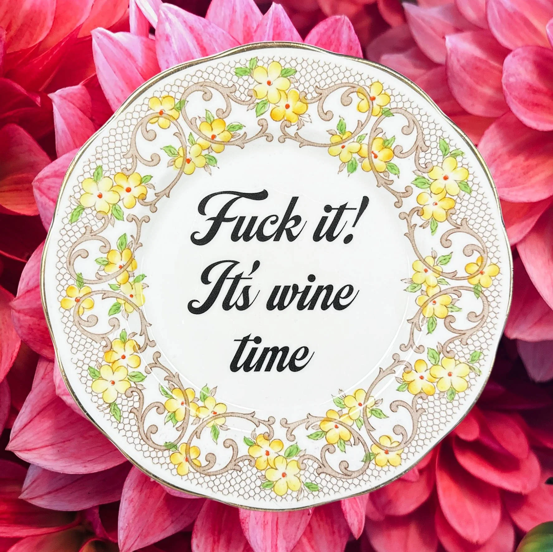 A vintage decorative wall plate with a yellow floral design and the words “fuck it! It’s wine time” in black writing