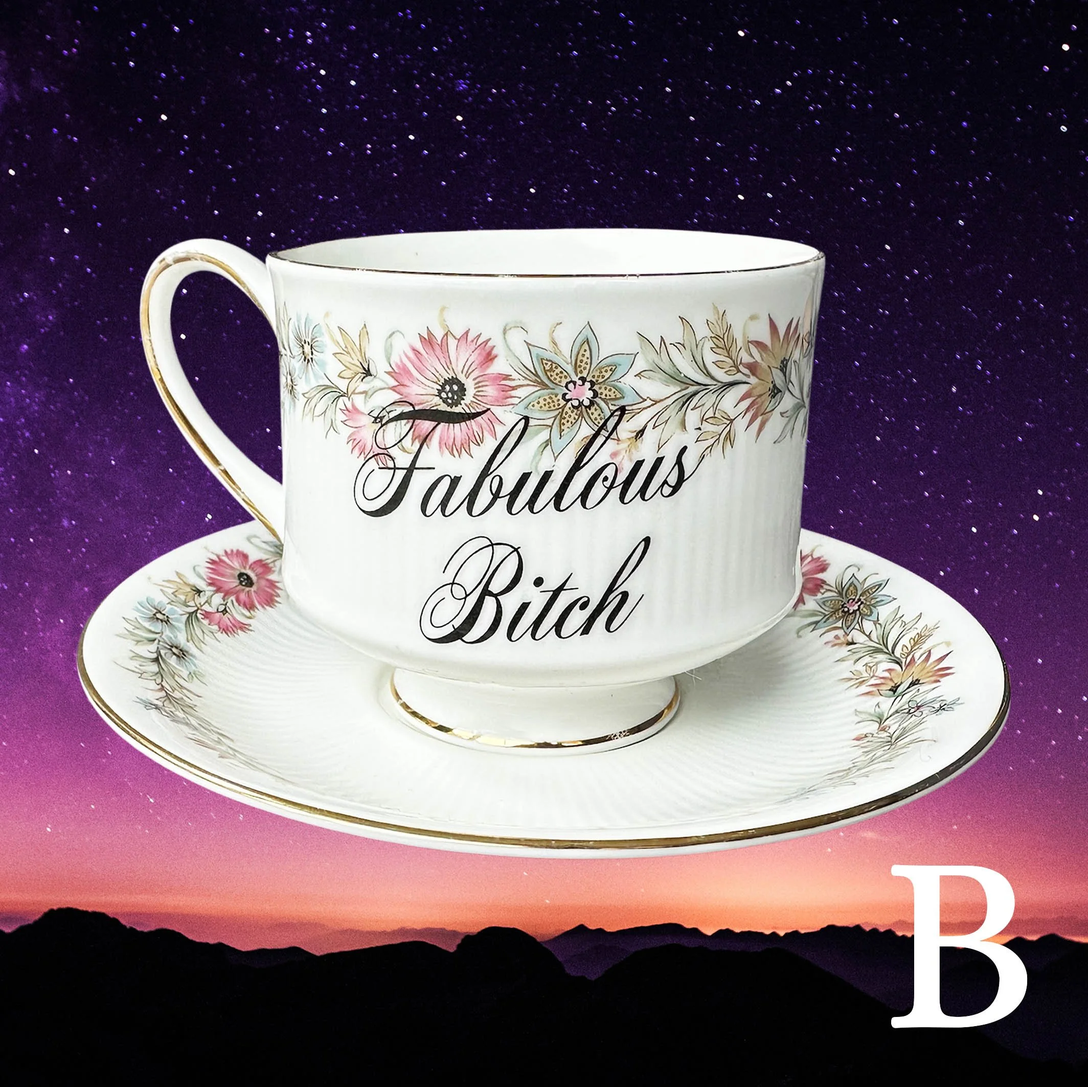 A vintage tea cup and saucer with a pink and blue floral design and the words “fabulous bitch” in black cursive writing