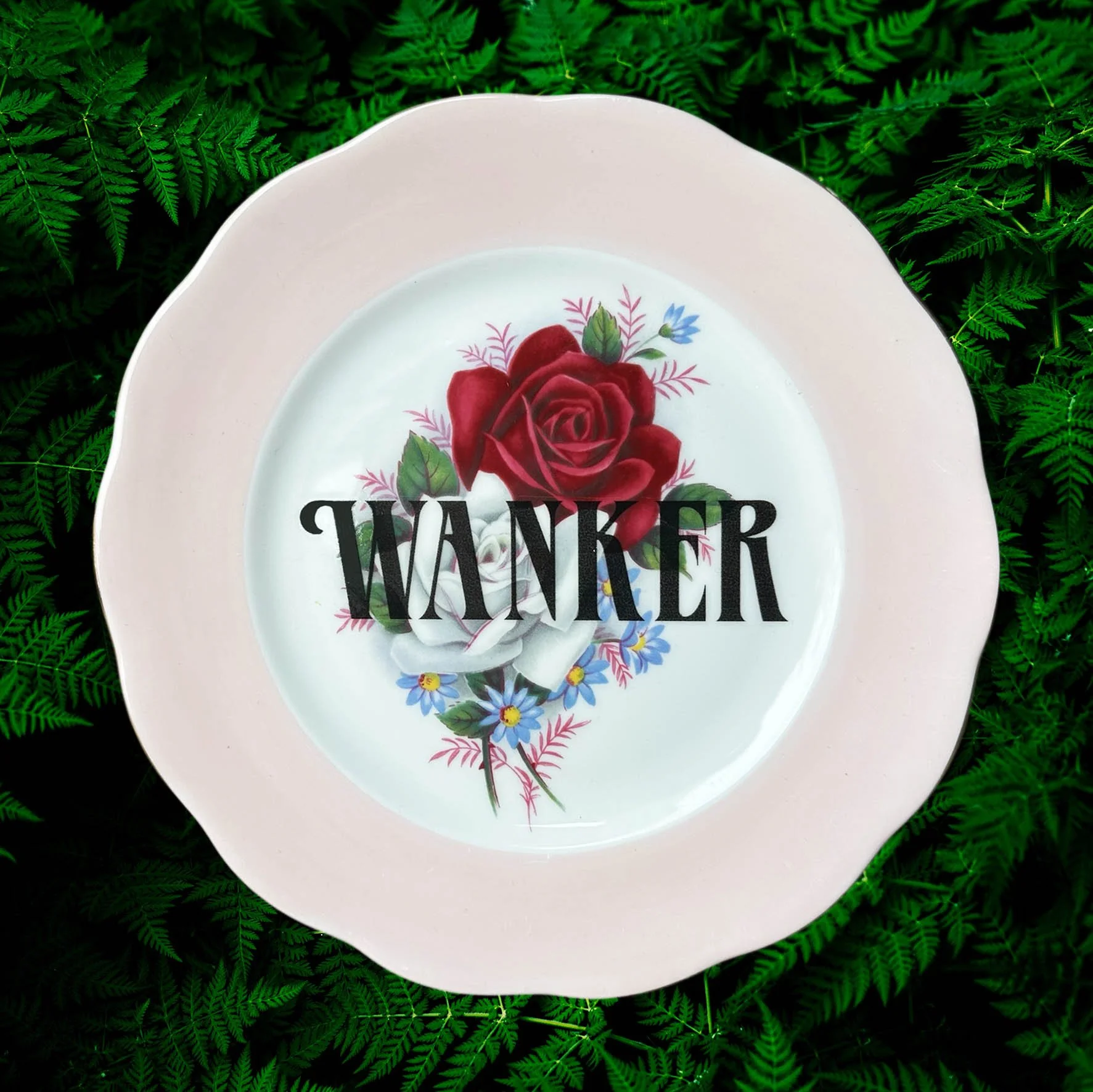 A vintage decorative wall plate with a red rose design and the word “wanker” in black lettering