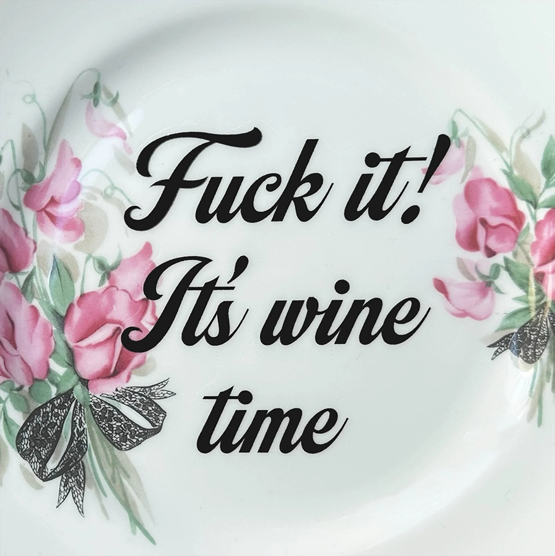 A vintage decorative wall plate with a pink floral design and the words “fuck it! It’s wine time” in black writing