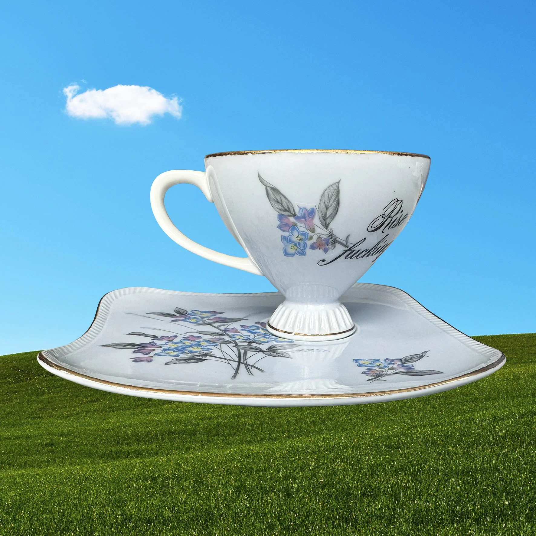A blue tea cup and saucer set with a blue floral design and the words “rise and fucking shine” in black cursive writing