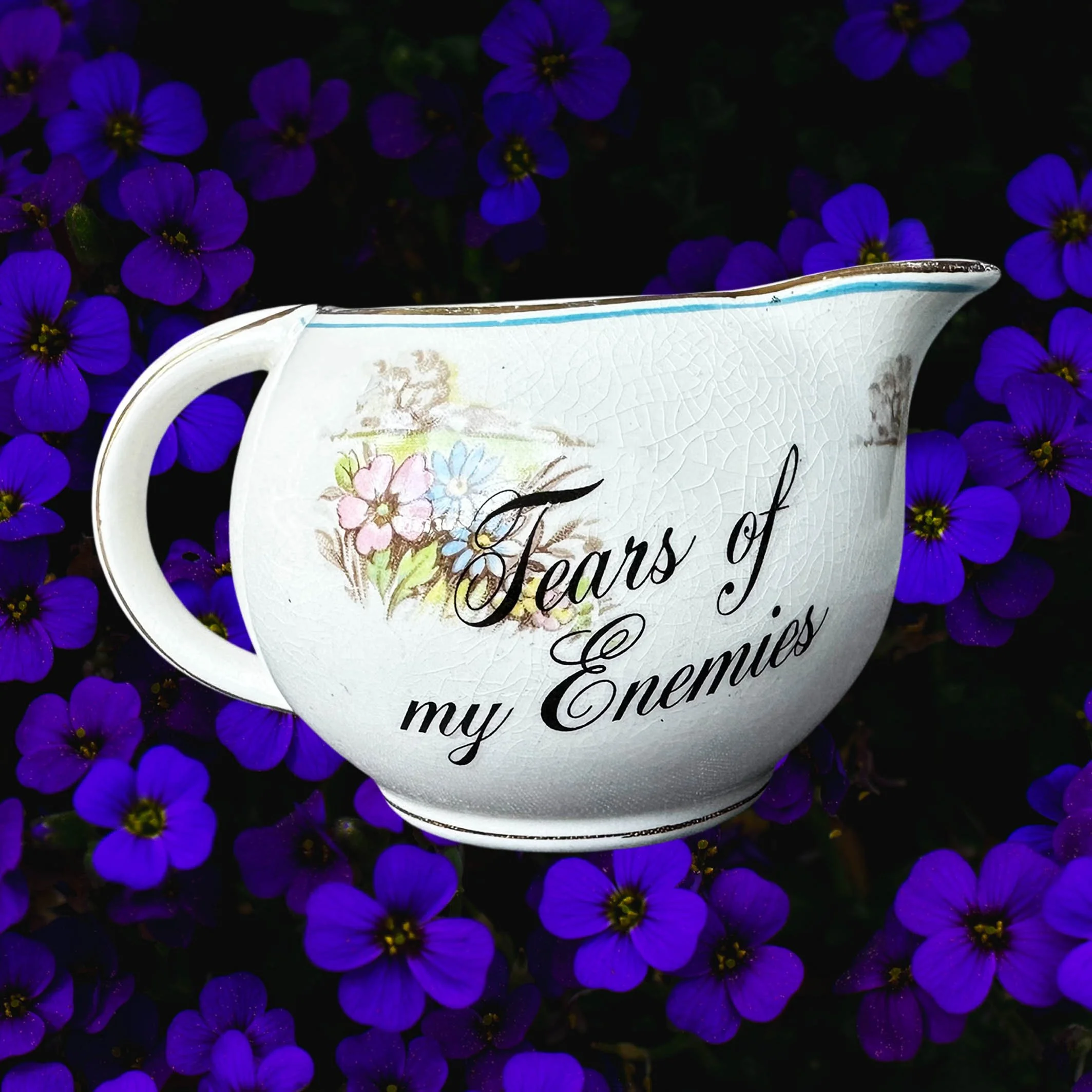 A cream coloured vintage milk jug with a floral design, the words “tears of my enemies” in black cursive writing