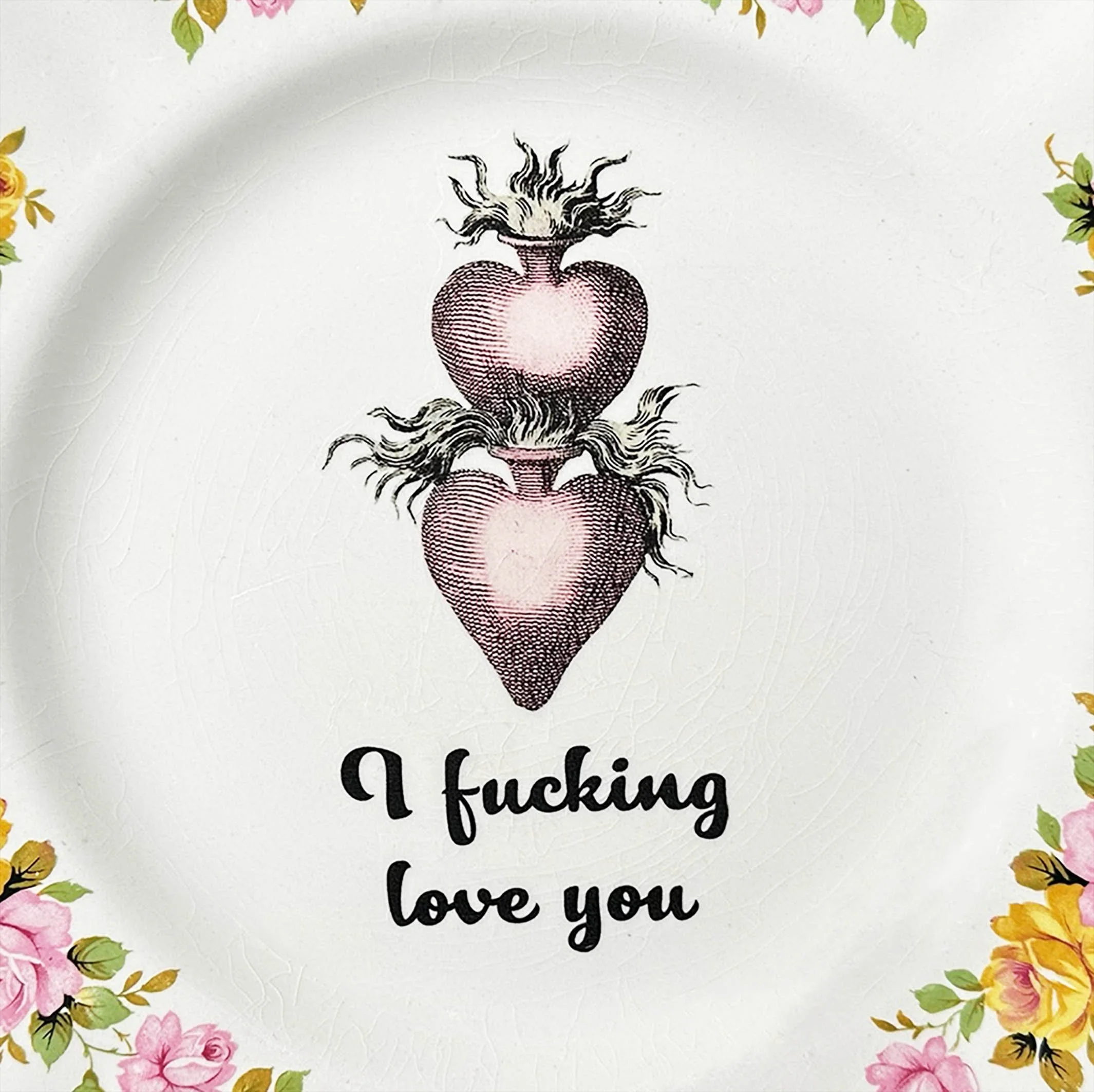 A vintage plate with a pink and yellow rose design picture two stacked hearts, with the words “I fucking love you” in black cursive writing
