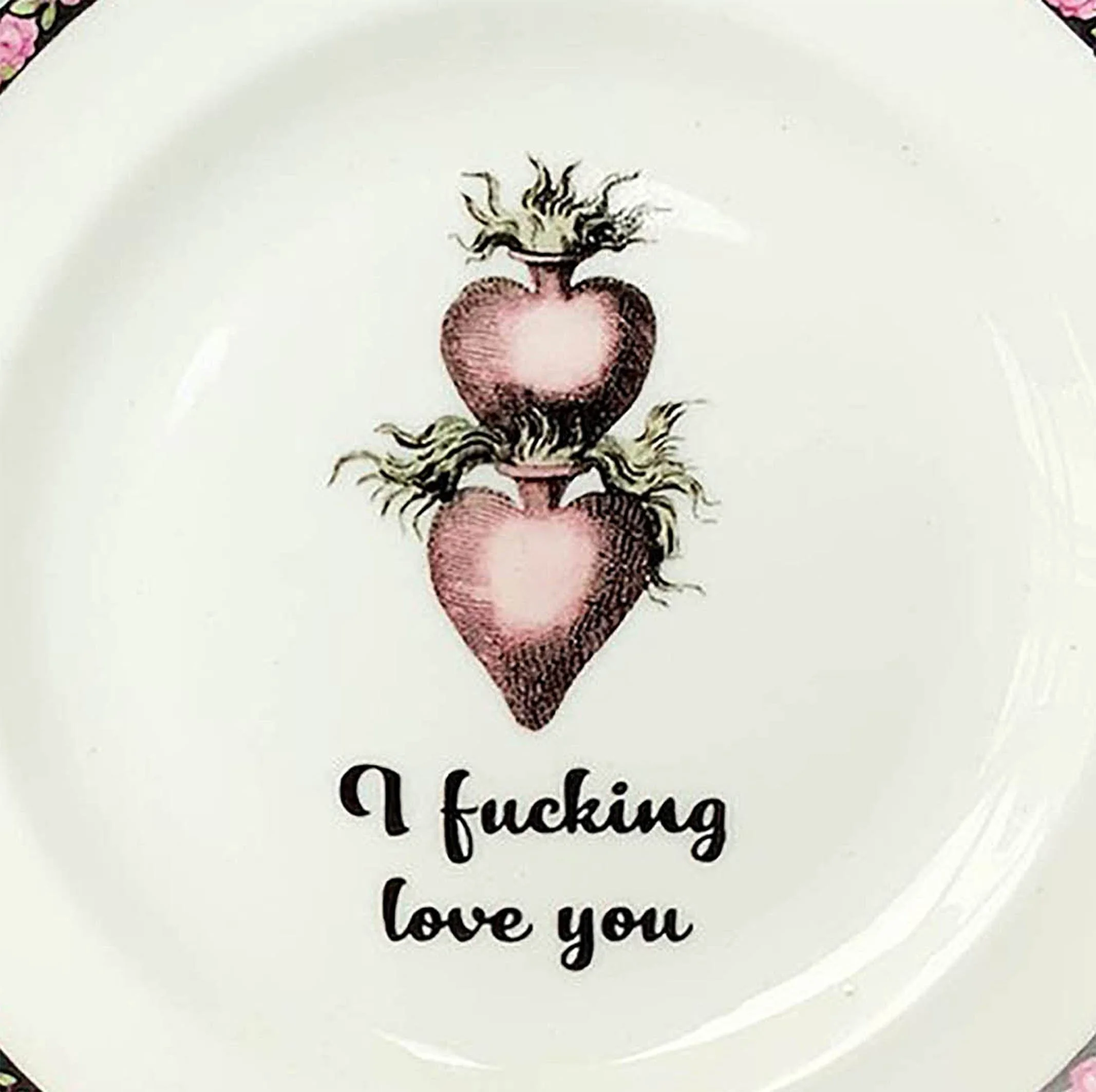 A vintage plate with a pink floral border design and a picture of two stacked hearts, and the words “I fucking love you” in black cursive writing