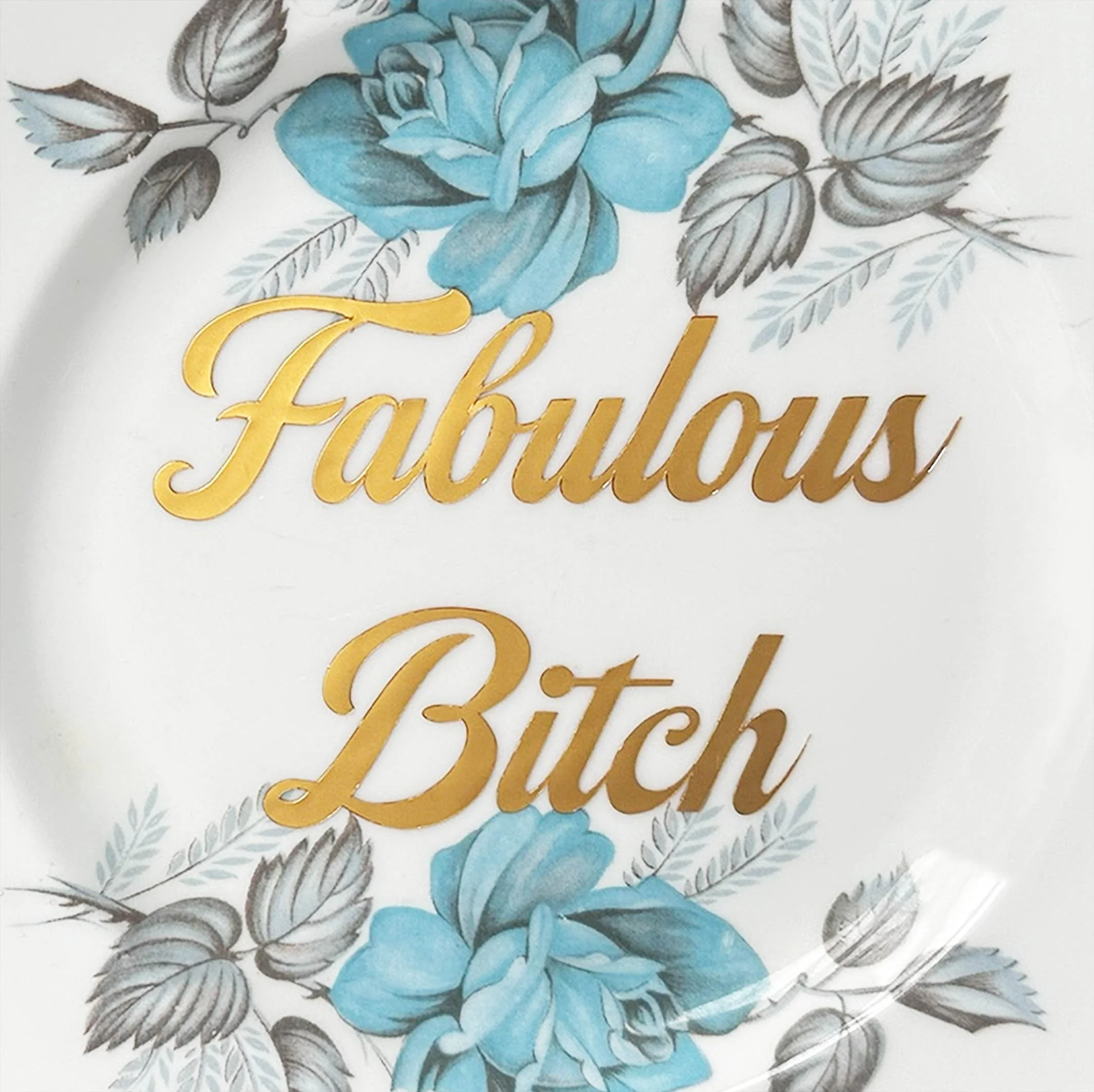 A vintage decorative wall plate with a blue floral design and the words “fabulous bitch” in gold lettering