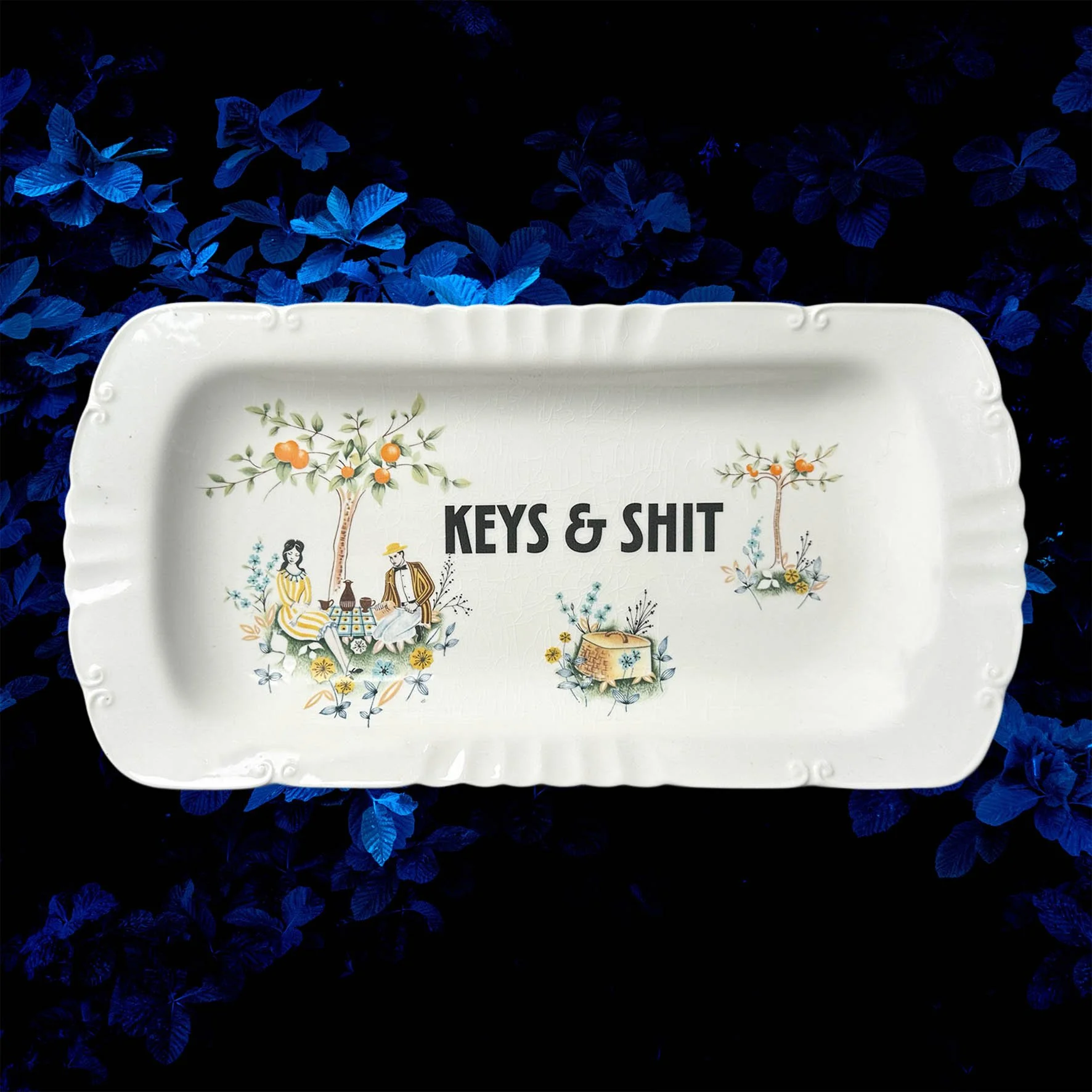A vintage trinket dish with a picnic scene and the words “keys & shit” in black lettering