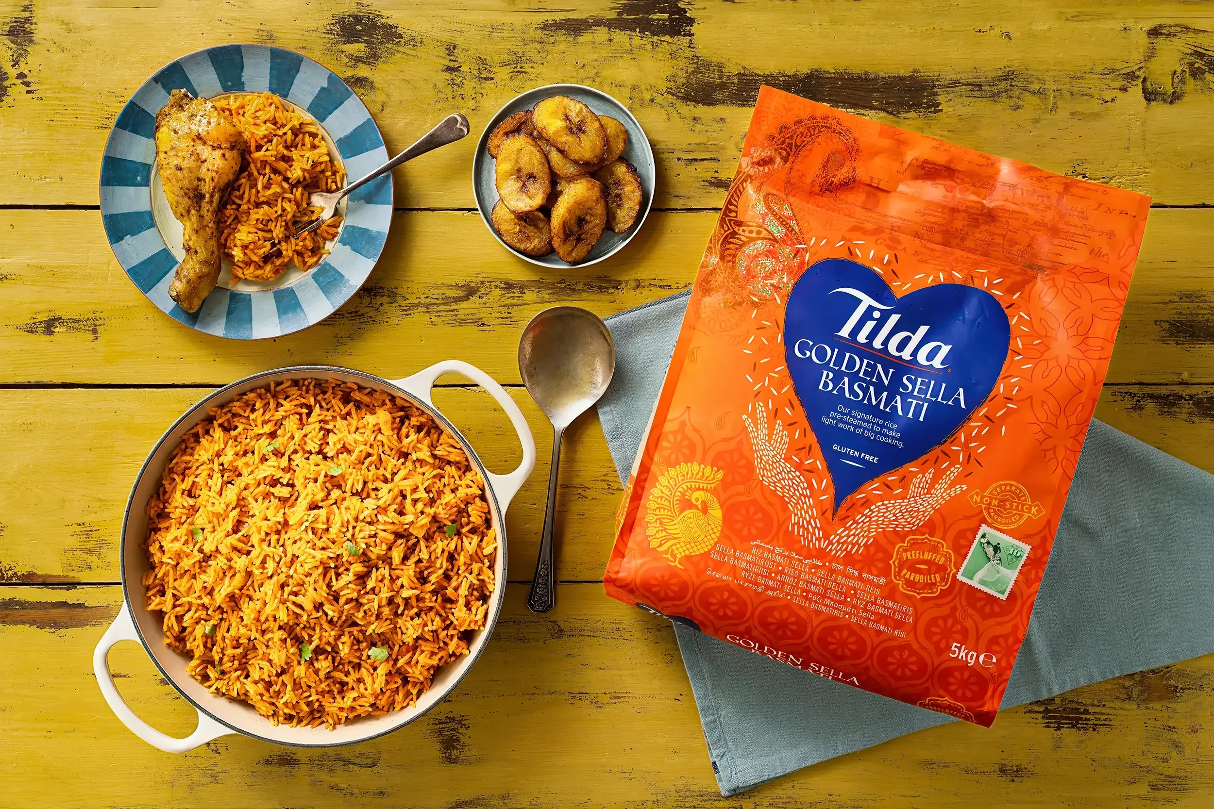 A large pot of cooked orange rice, a plate with a piece of chicken leg and more rice, a bowl of fried plantains, and a package of Tilda Golden Sella Basmati rice on a yellow table with a blue cloth underneath the rice package.