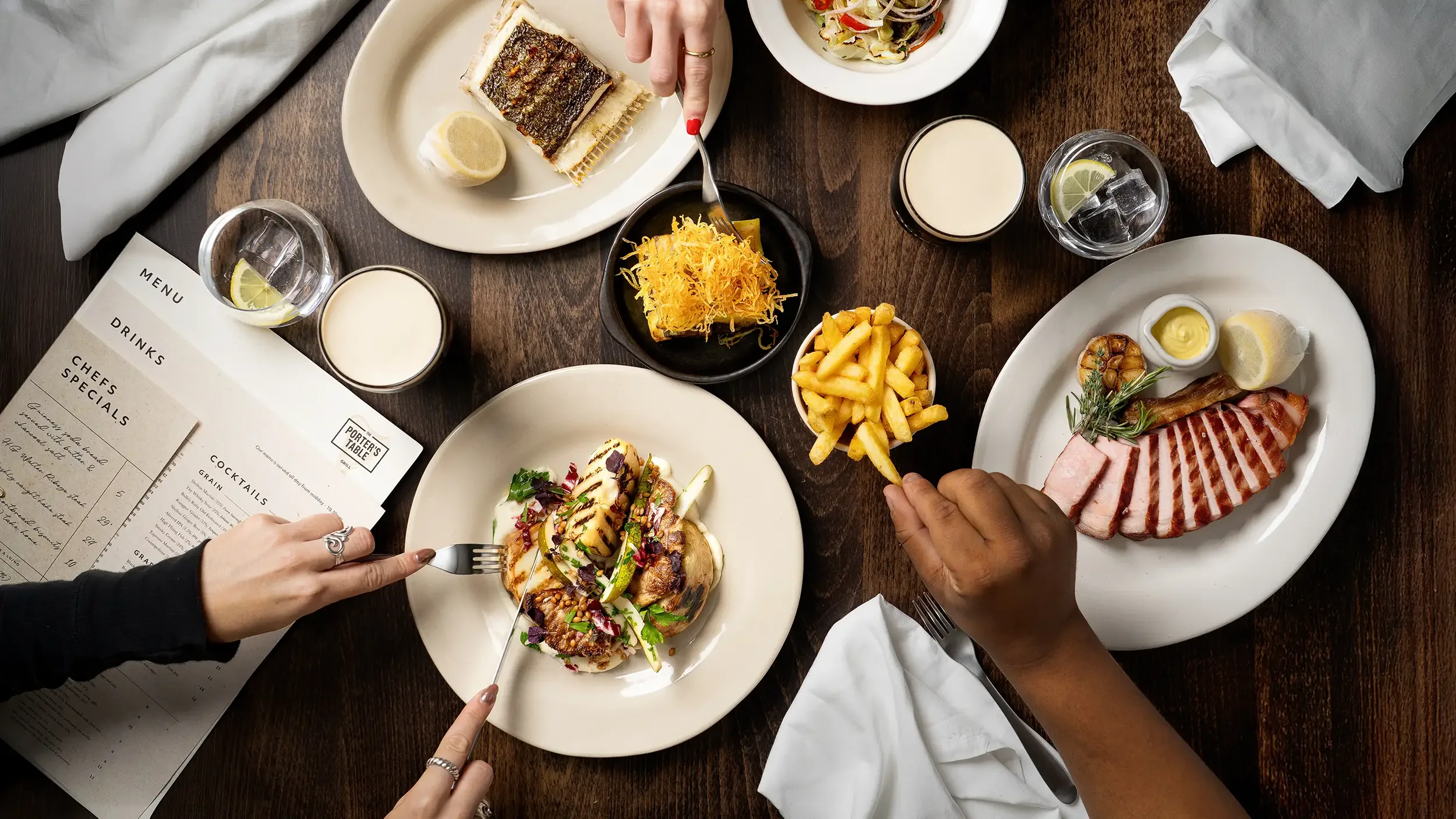 A top-down view of a dinner table with various dishes, including grilled meat, fries, a seafood dish, a salad, and drinks. There are hands reaching for food and a menu on the table.