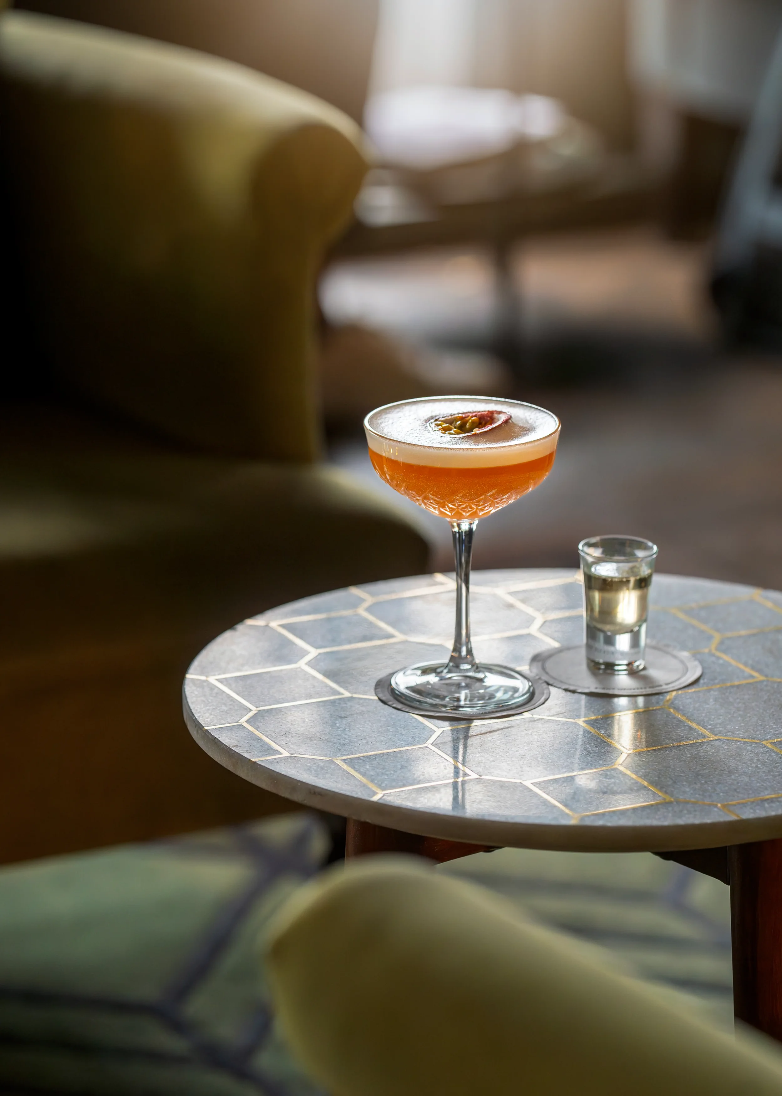 A cocktail glass sits on a round table with a hexagonal tile pattern, with a small shot glass next to it. The background is softly blurred with warm lighting.