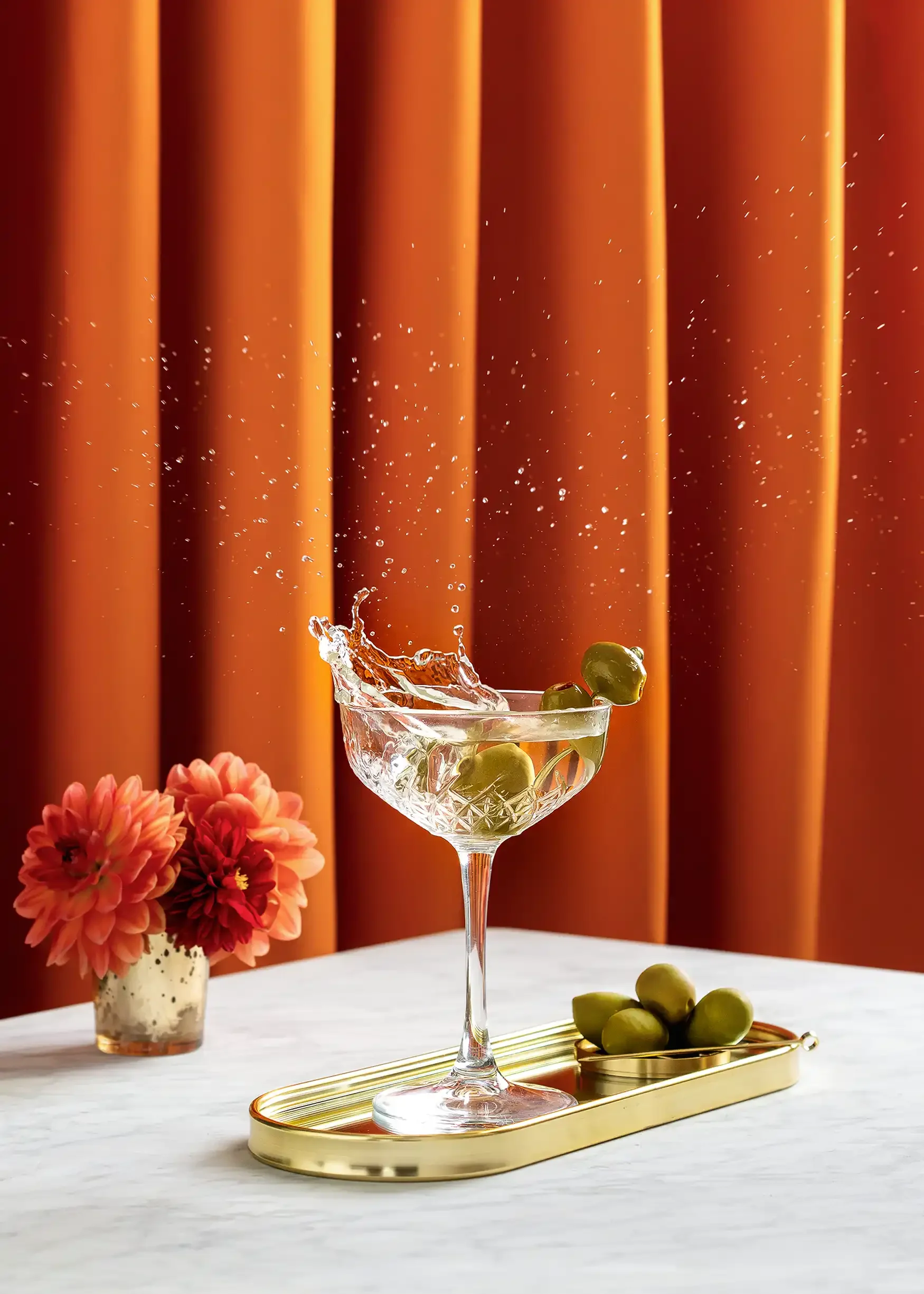A crystal glass with green olives inside, placed on a gold tray on a white table. There is a vase of pink and orange flowers in the background, and a bunch of green olives on the table. The backdrop features orange curtains.
