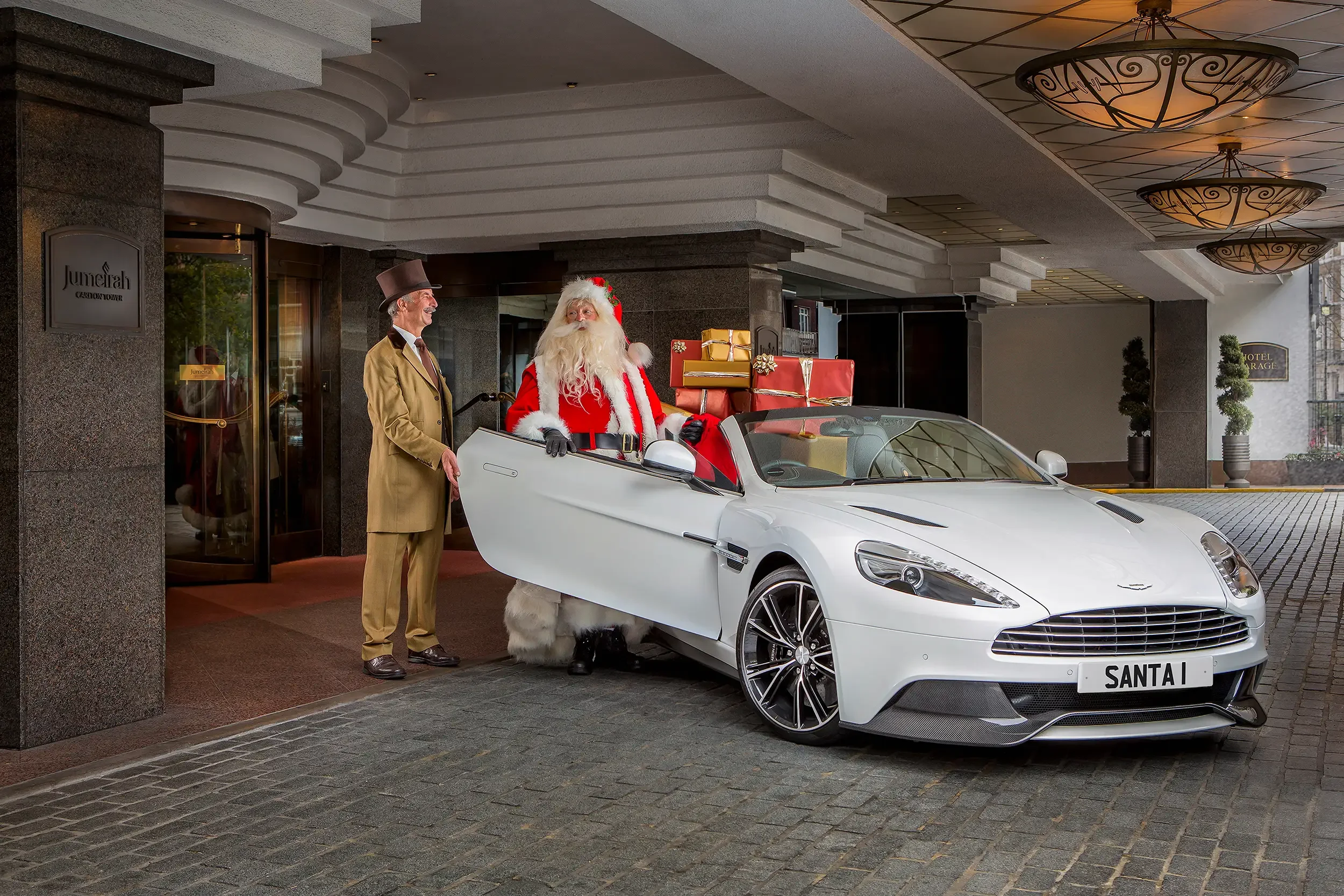 Santa Claus sitting in a white luxury sports car with a license plate that reads 'SANTA1,' surrounded by wrapped Christmas presents, outside a hotel lobby. An assistant in a beige suit with a top hat is standing next to the car.