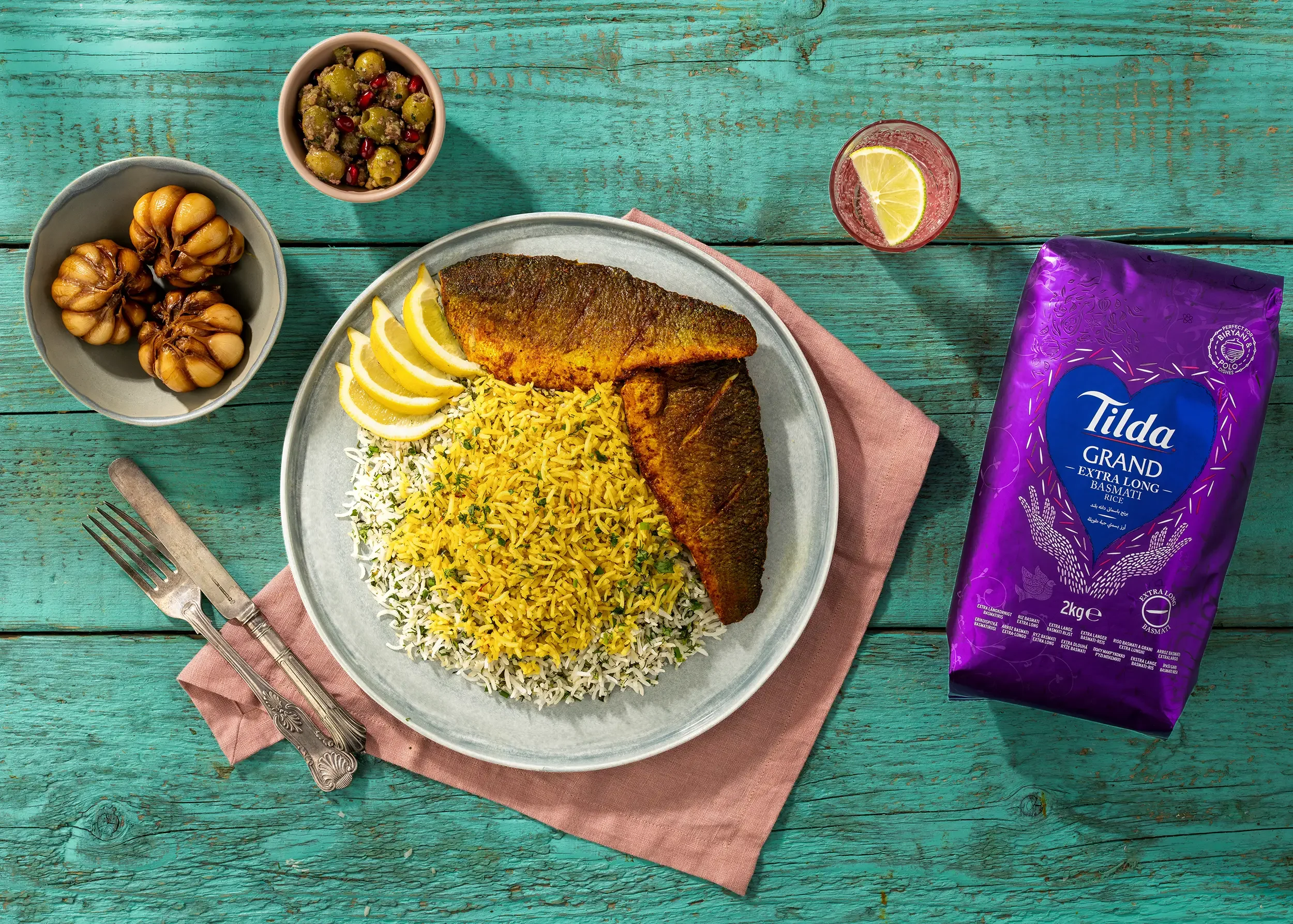 Plate of rice with fried fish, lemon wedges, and side items including green olives with pomegranate seeds, browned garlic cloves, and a red beverage with lemon slice in a glass, all on a turquoise wooden table with a pink napkin and vintage fork and 