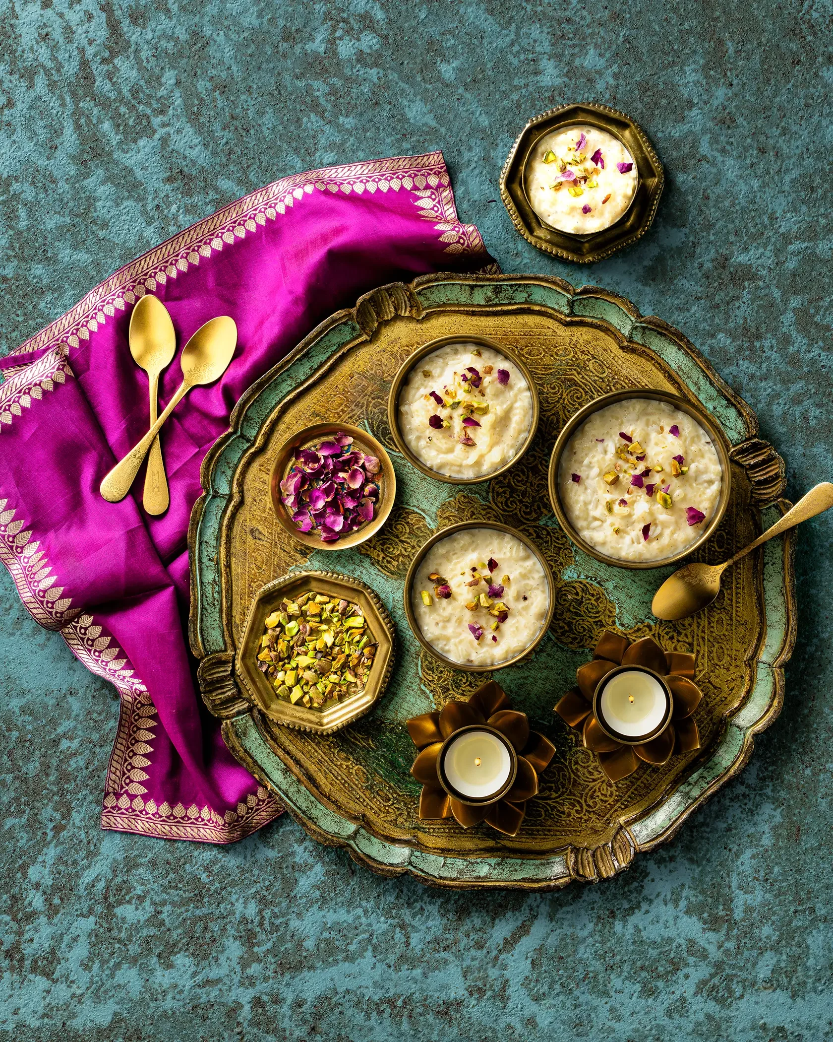 Traditional Indian desserts served in bowls on a decorative tray, accompanied by a pink fabric with gold accents, gold spoons, rose petals, chopped pistachios, and candles.