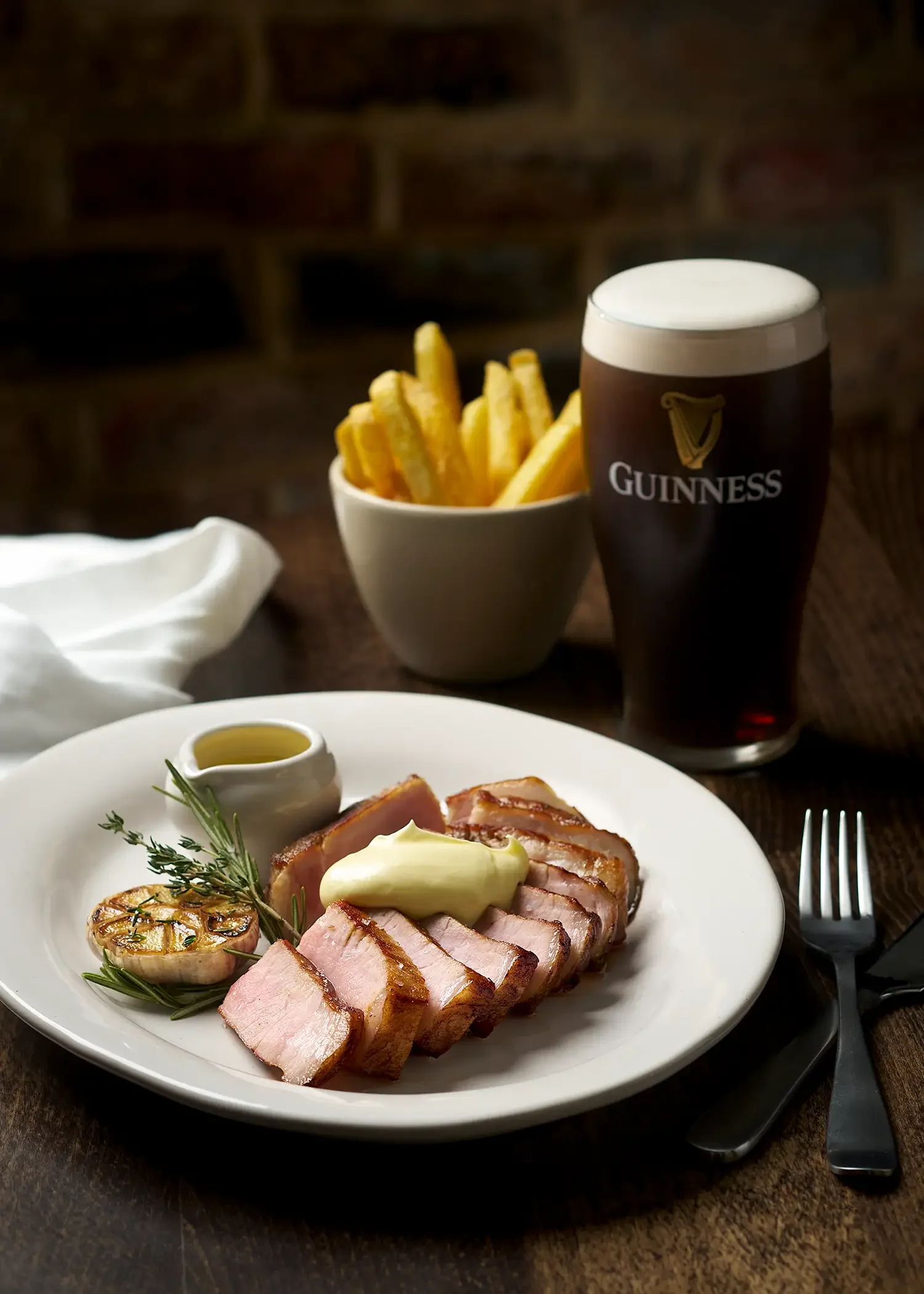 Plate of sliced meat with garnishes, garlic, and herbs, alongside a small dish of sauce, a bowl of French fries, and a pint of Guinness beer on a wooden table.