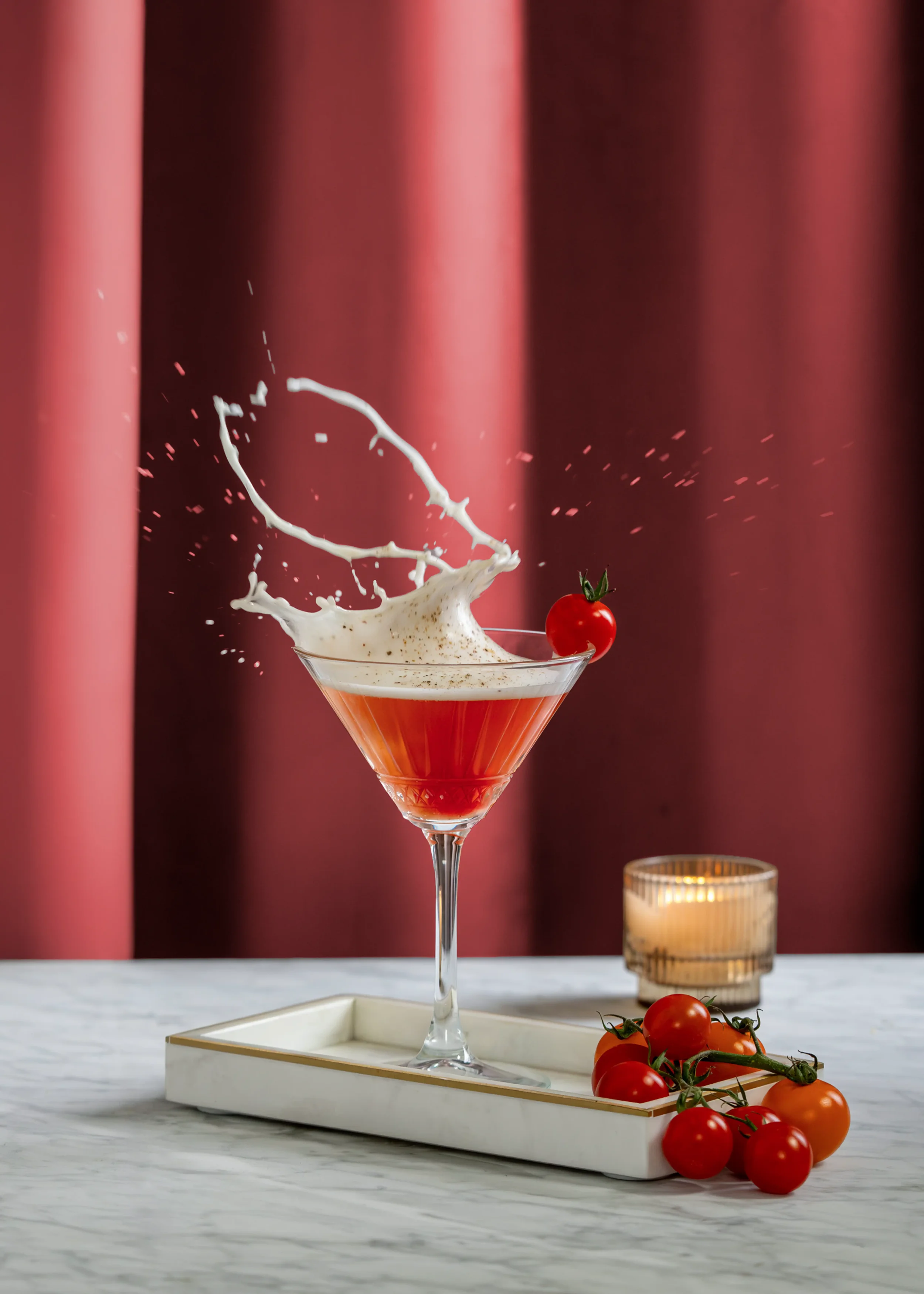 A pink cocktail glass with a cherry on top, with cream splashing into the drink, set on a white tray with cherry tomatoes and a glass candle holder in the background, against a deep red curtain.