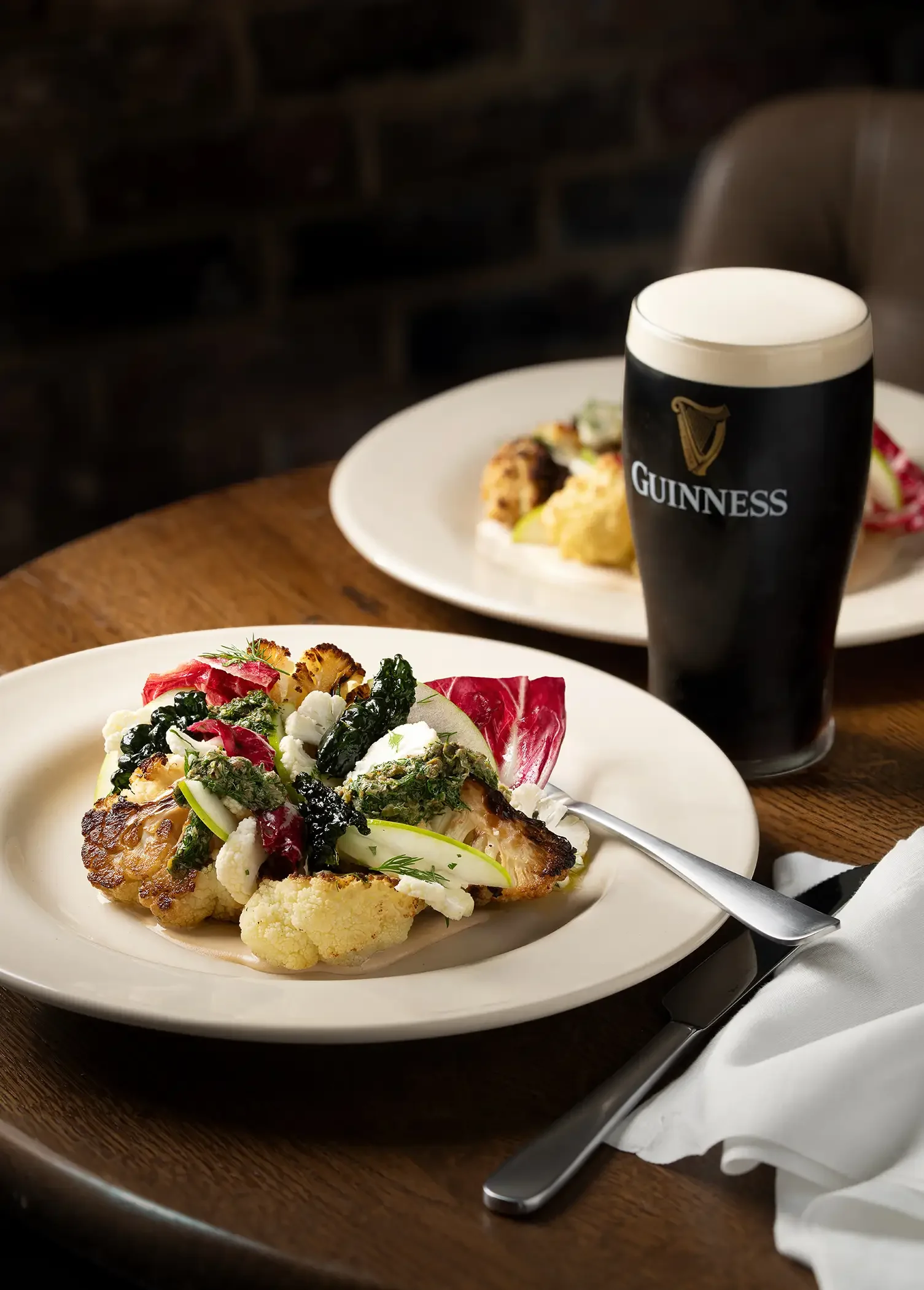 A plate of roasted vegetables, including cauliflower, broccoli, carrots, and zucchini, with greens and herbs, served on a white plate next to a knife and white napkin. In the background, there is another plate of food and a pint of Guinness beer on a