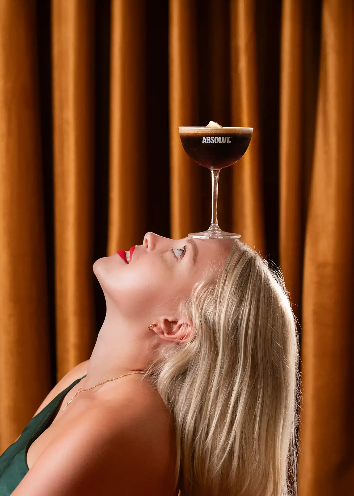 A woman with blonde hair lying on her back, with a cocktail glass balanced on her forehead. The cocktail is dark-colored with a frothy top and the word 'ABSOLUT' visible on the glass, against a backdrop of brown curtains.