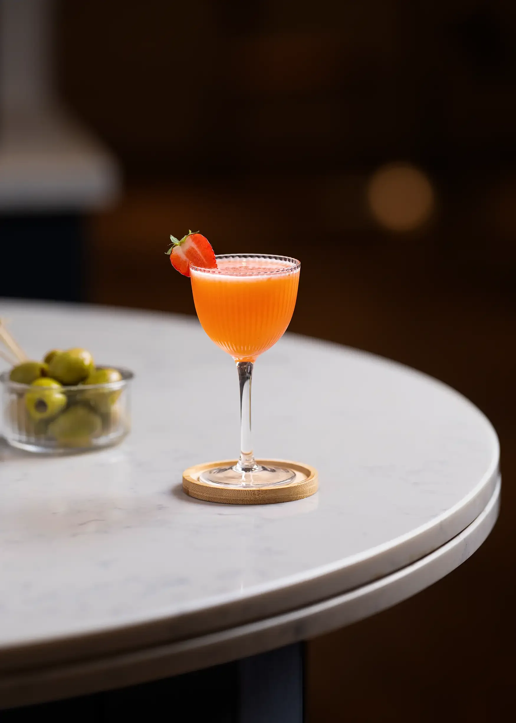 A pink cocktail in a tall glass with a strawberry slice garnish on the rim, placed on a small wooden coaster on a white table.