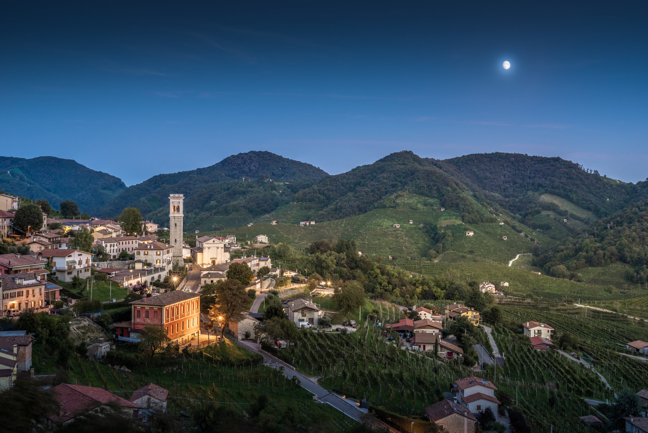 Paesaggio montano con un piccolo villaggio, chiesa con torre campanaria, case, vigneti e sentieri, sotto un cielo notturno con la luna