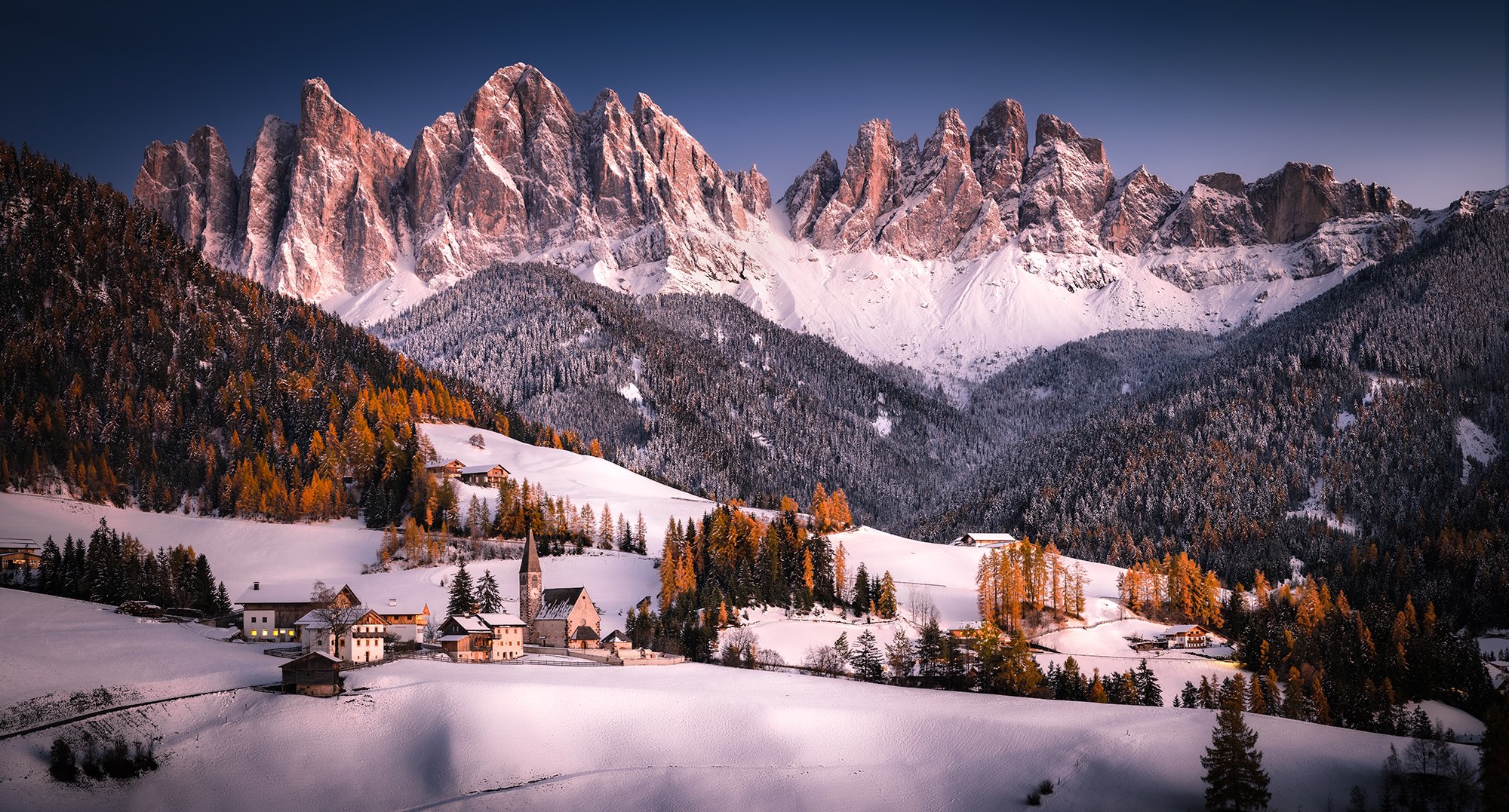 Paesaggio montano innevato con un villaggio, chiese e boschi, sullo sfondo le Dolomiti all'alba o al tramonto.