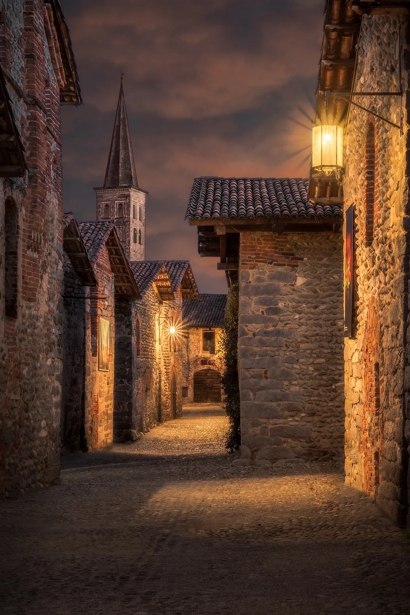 Una stretta strada di ciottoli in un villaggio medievale con case in pietra e tetti di tegole, illuminata da lampioni a luce calda, con un campanile in cima di un edificio religioso visibile sullo sfondo, al tramonto o sera.