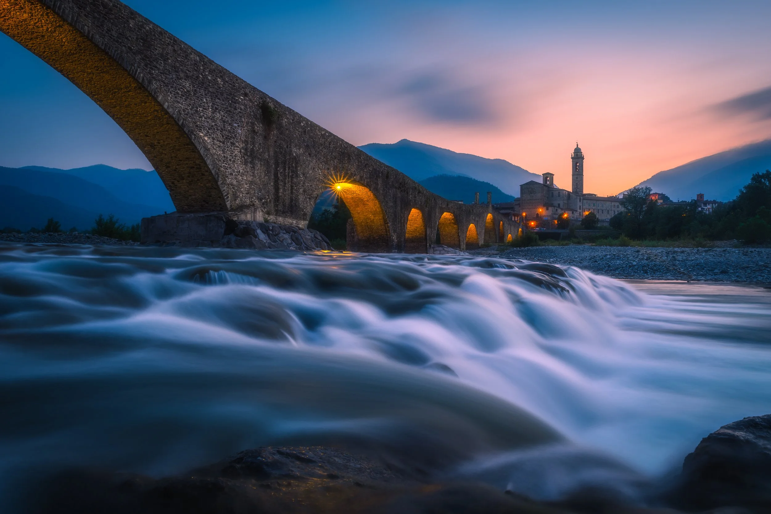 Ponte di pietra su un fiume con acqua in movimento, al tramonto, con edifici e montagne sullo sfondo in Italia.