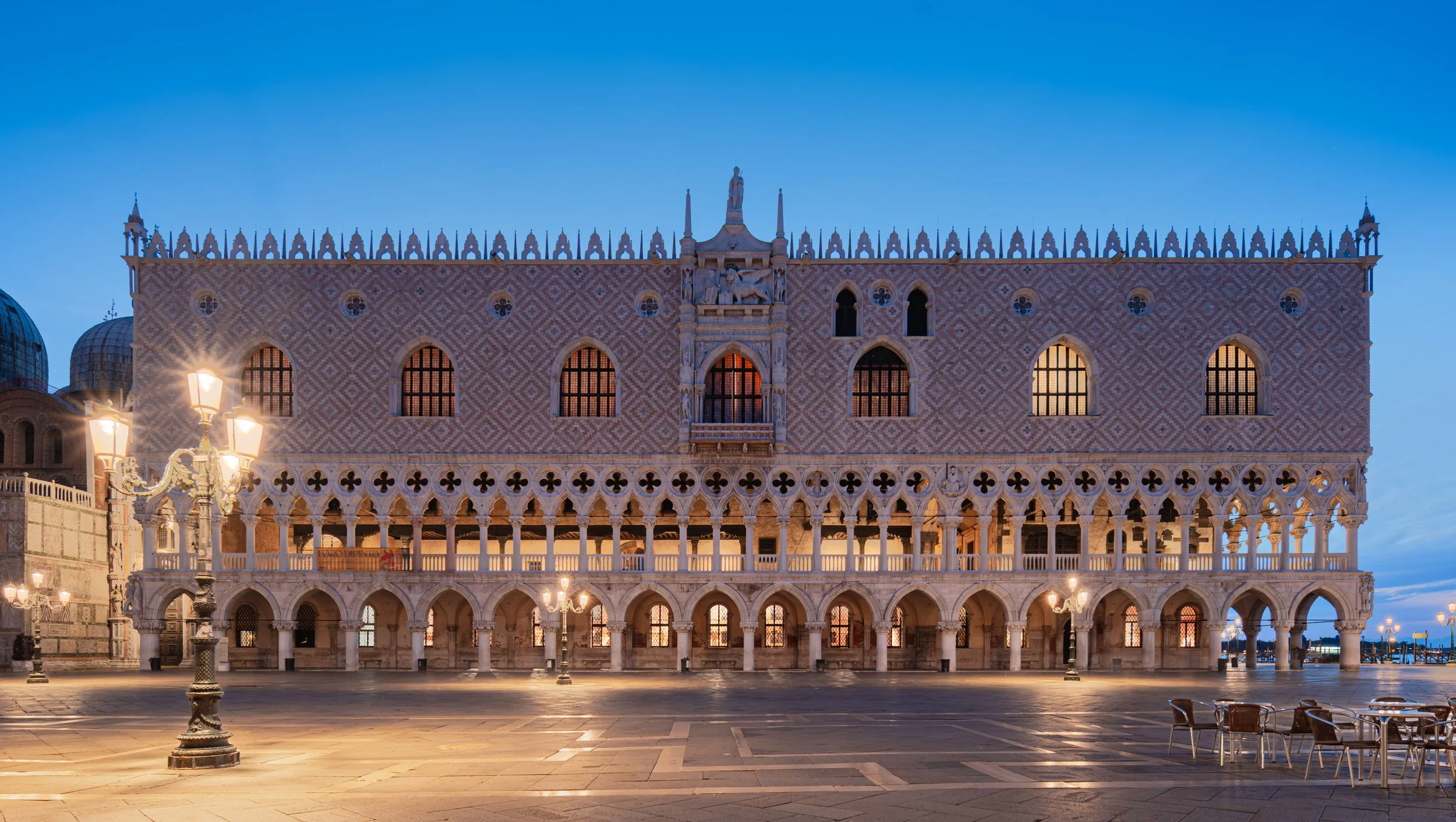 Il Palazzo Ducale di Venezia vista di sera con le luci accese e un cielo blu. Davanti ci sono lampioni elegantemente decorati e alcune sedie.