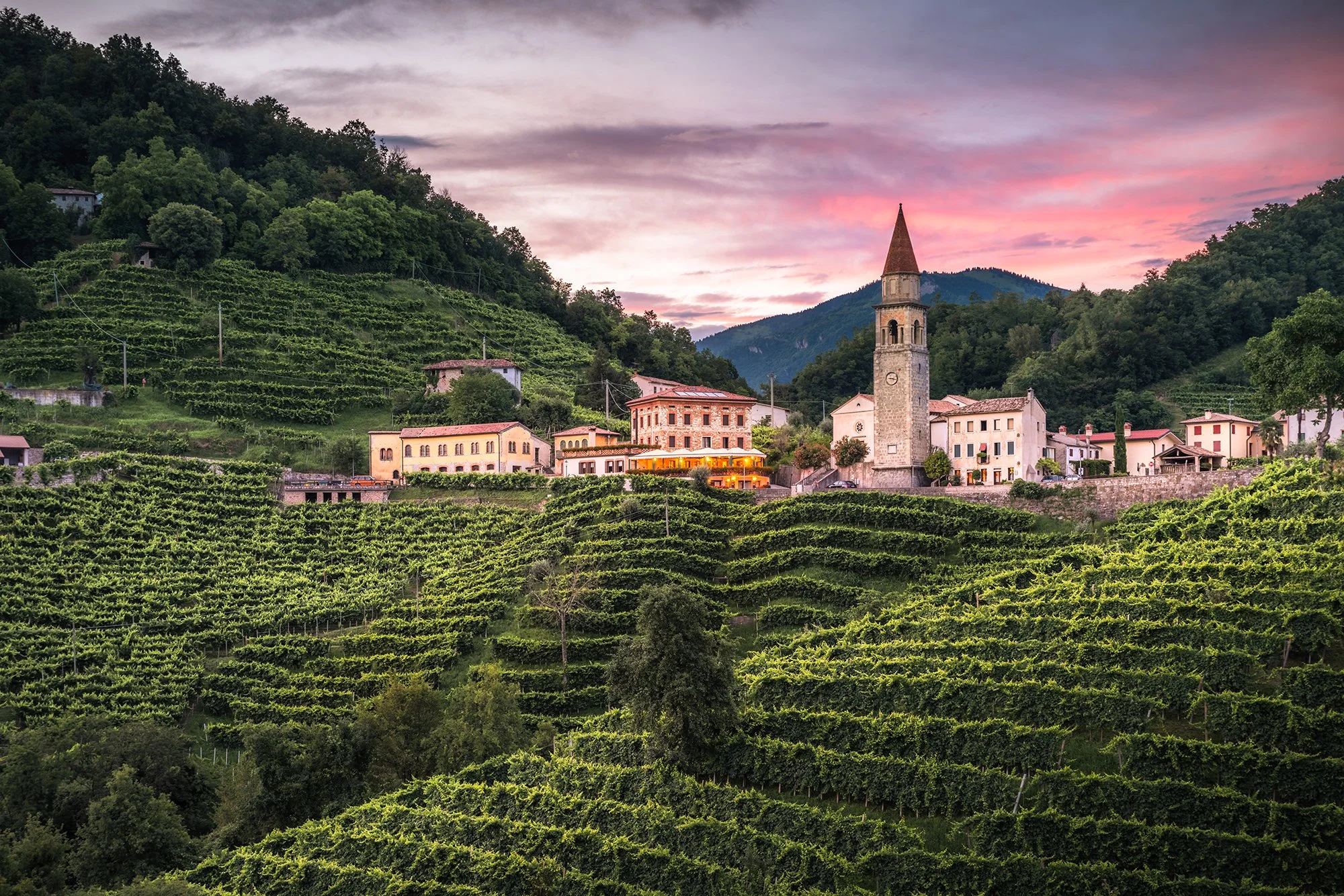 Paesaggio di un paese di montagna con case, chiesa con torre e vigna verdeggiante durante il tramonto.