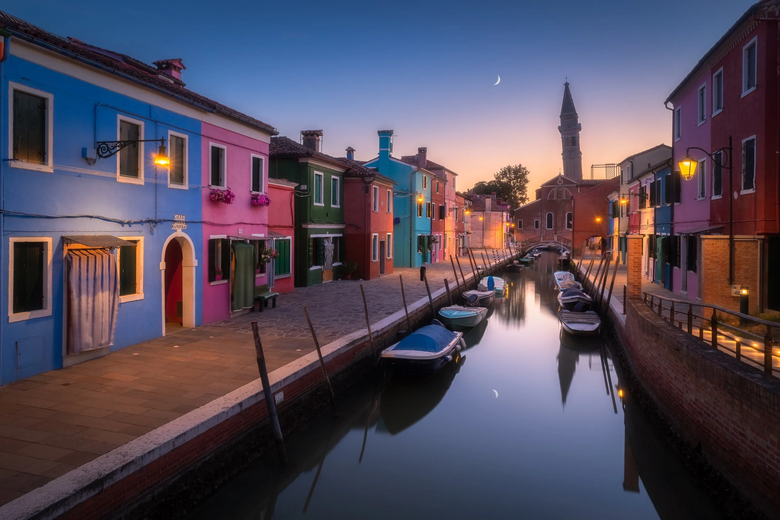 Canal con barche ormeggiate accanto a case colorate lungo un percorso ciclopedonale, al tramonto, con una chiesa e il cielo con la luna calante visibili sullo sfondo.
