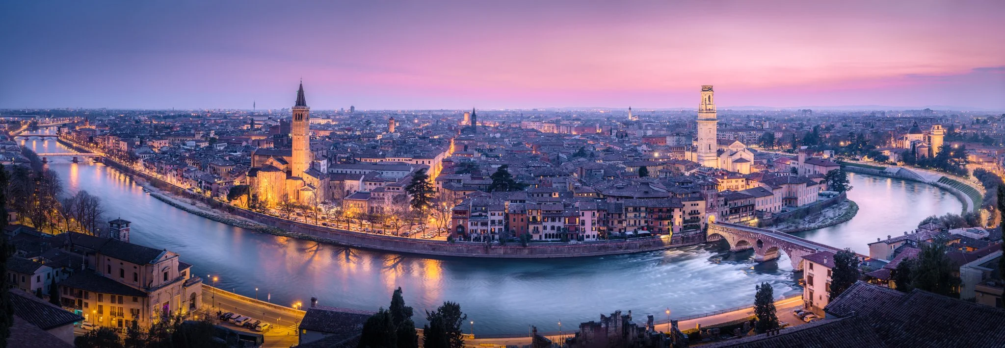 Panoramica di una città europea con fiume che attraversa, chiese con torri, edifici storici e un cielo al tramonto con sfumature rosa e viola.