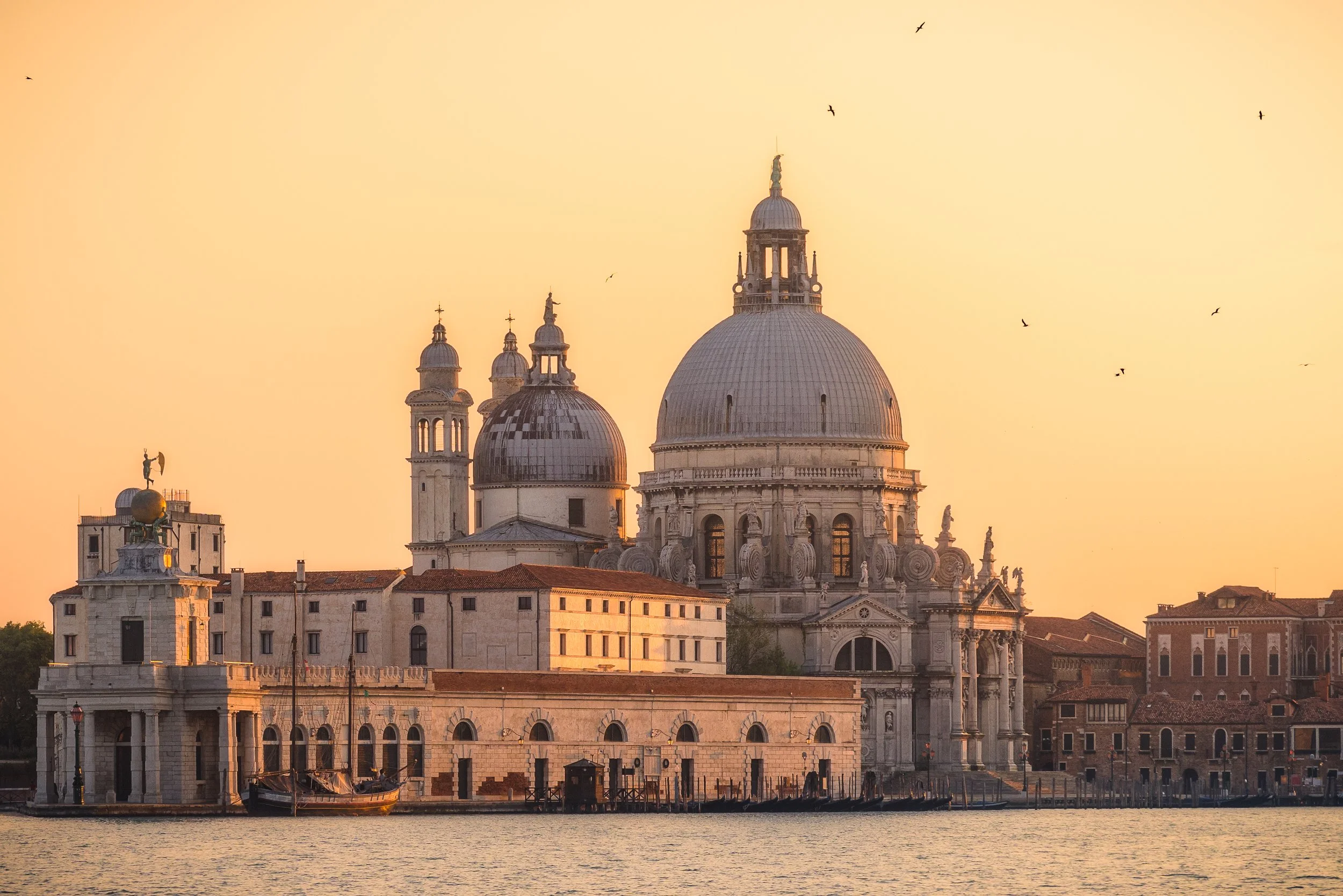 Veduta della Basilica di Santa Maria della Salute a Venezia durante il tramonto, con il cielo arancione e alcune barche sul fiume.