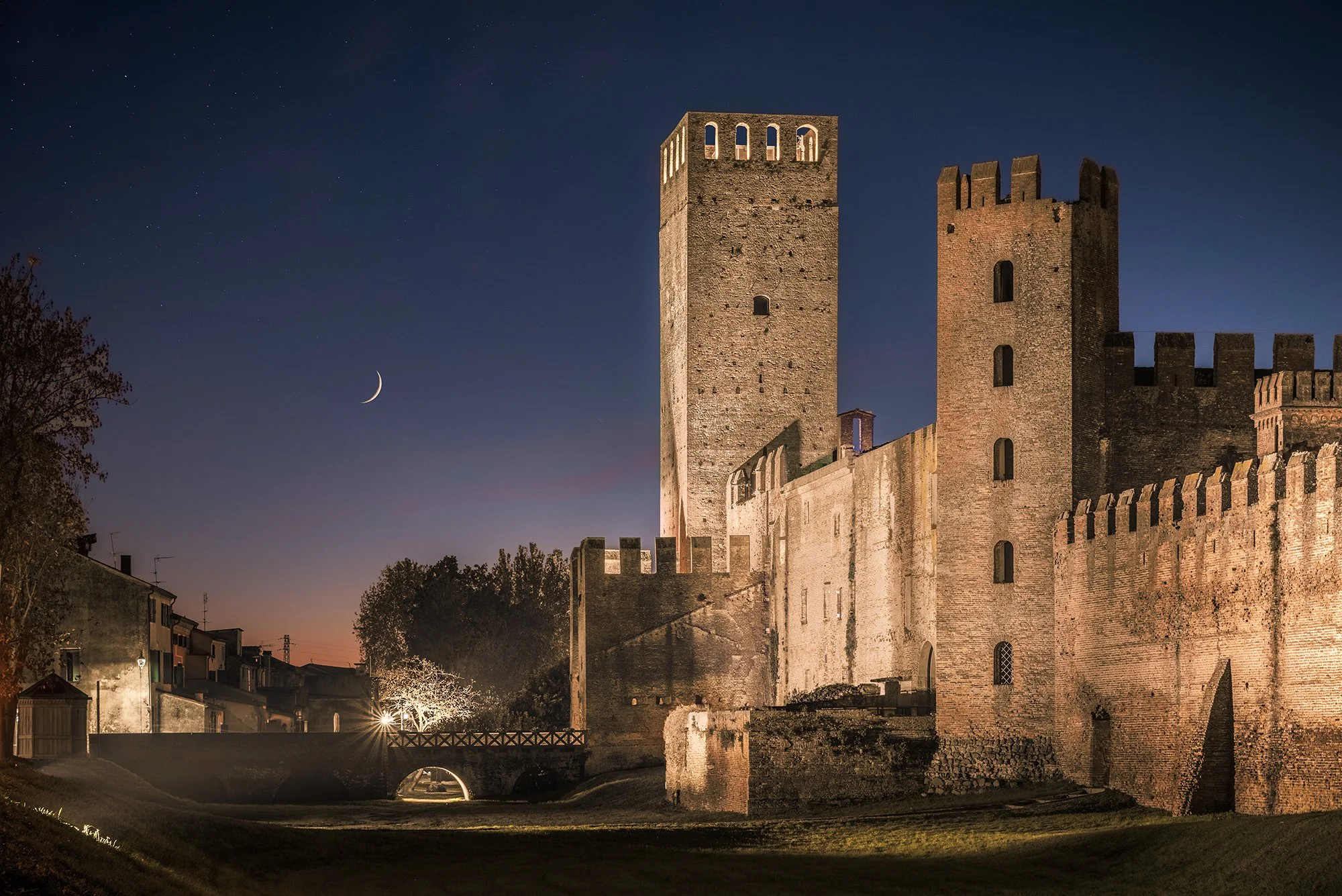 Castello medievale illuminato di notte con un cielo stellato e una luna crescente, alcune case e alberi sullo sfondo.