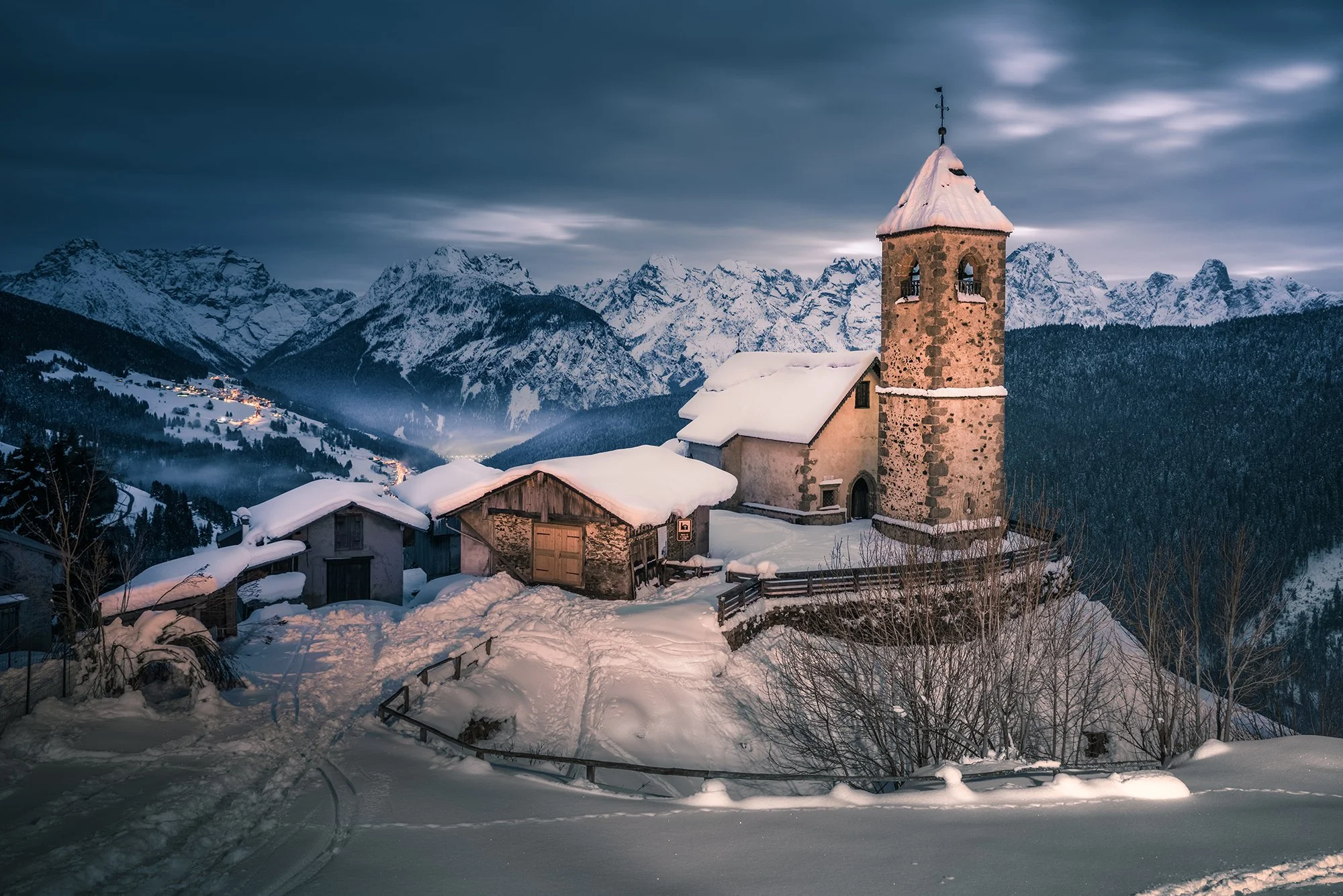 Paesaggio montano in inverno con una chiesa di pietra e case ricoperte di neve, montagne innevate sullo sfondo e cielo nuvoloso.