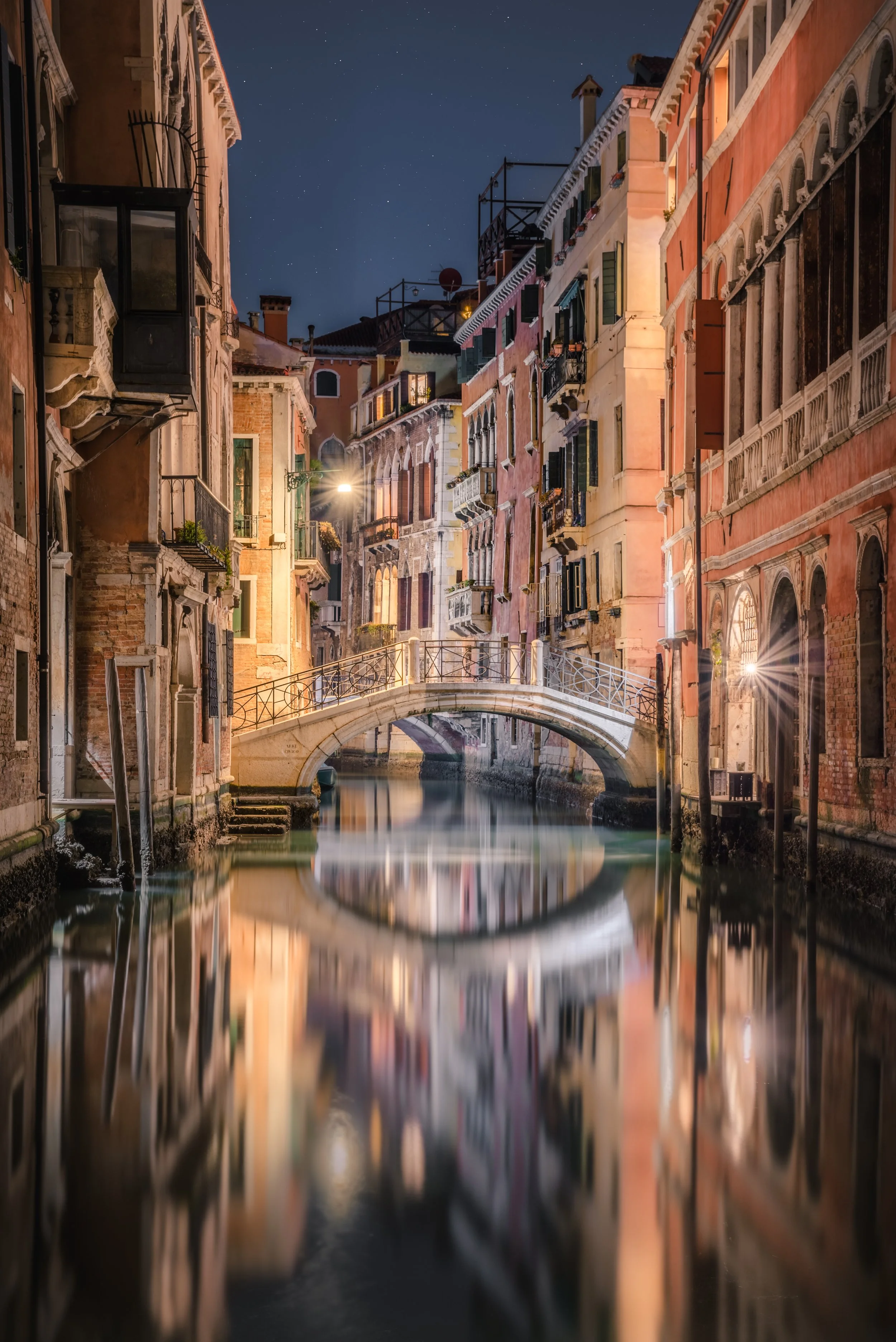 Vista notturna di un canale a Venezia con un ponte di pietra e edifici colorati ai lati, riflessi sull'acqua e il cielo stellato.