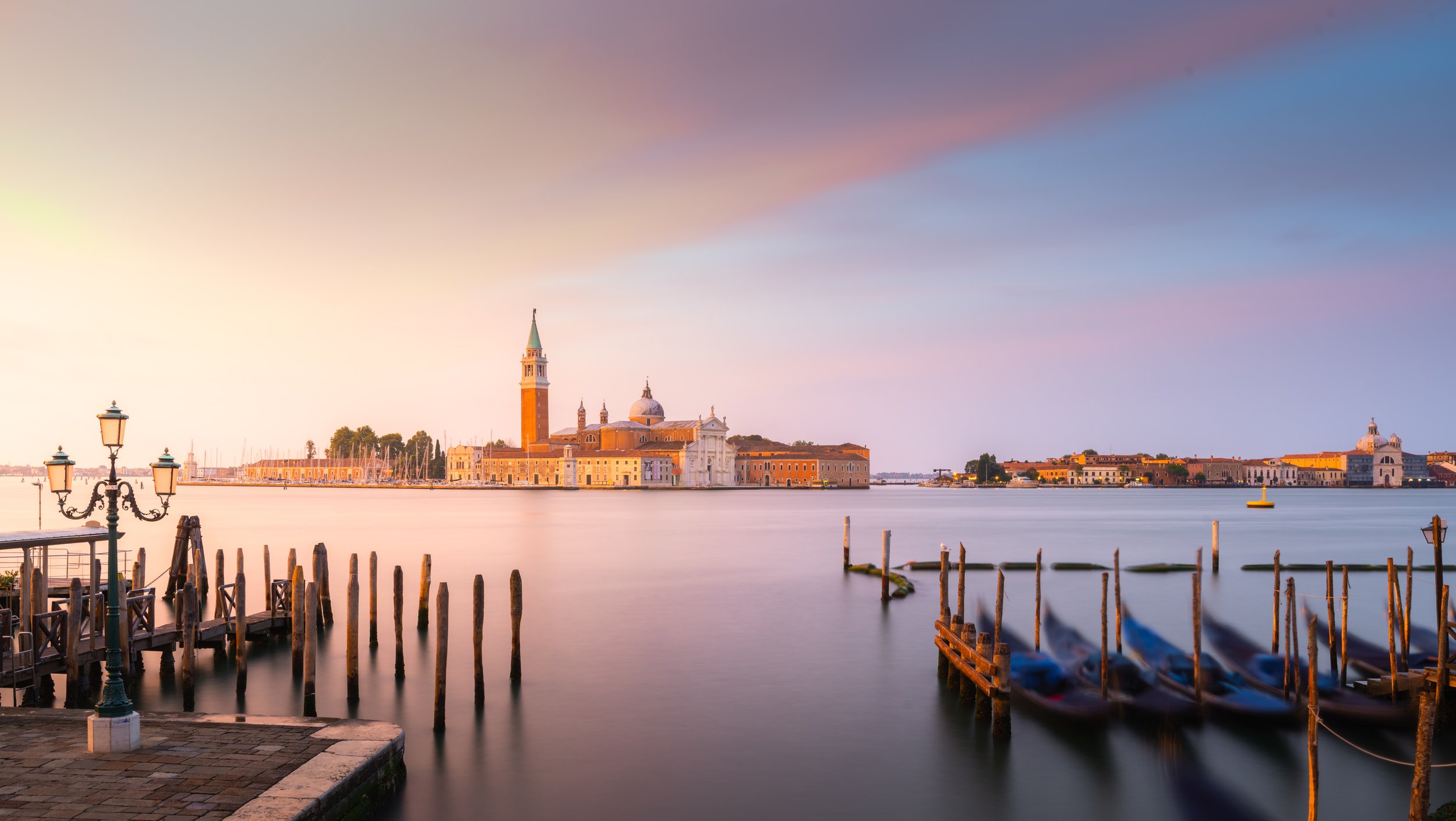 Vista panoramica di Venezia al tramonto, con gondole attraccate in primo piano, il campanile di San Marco e la basilica sullo sfondo, cielo colorato di rosa e viola.