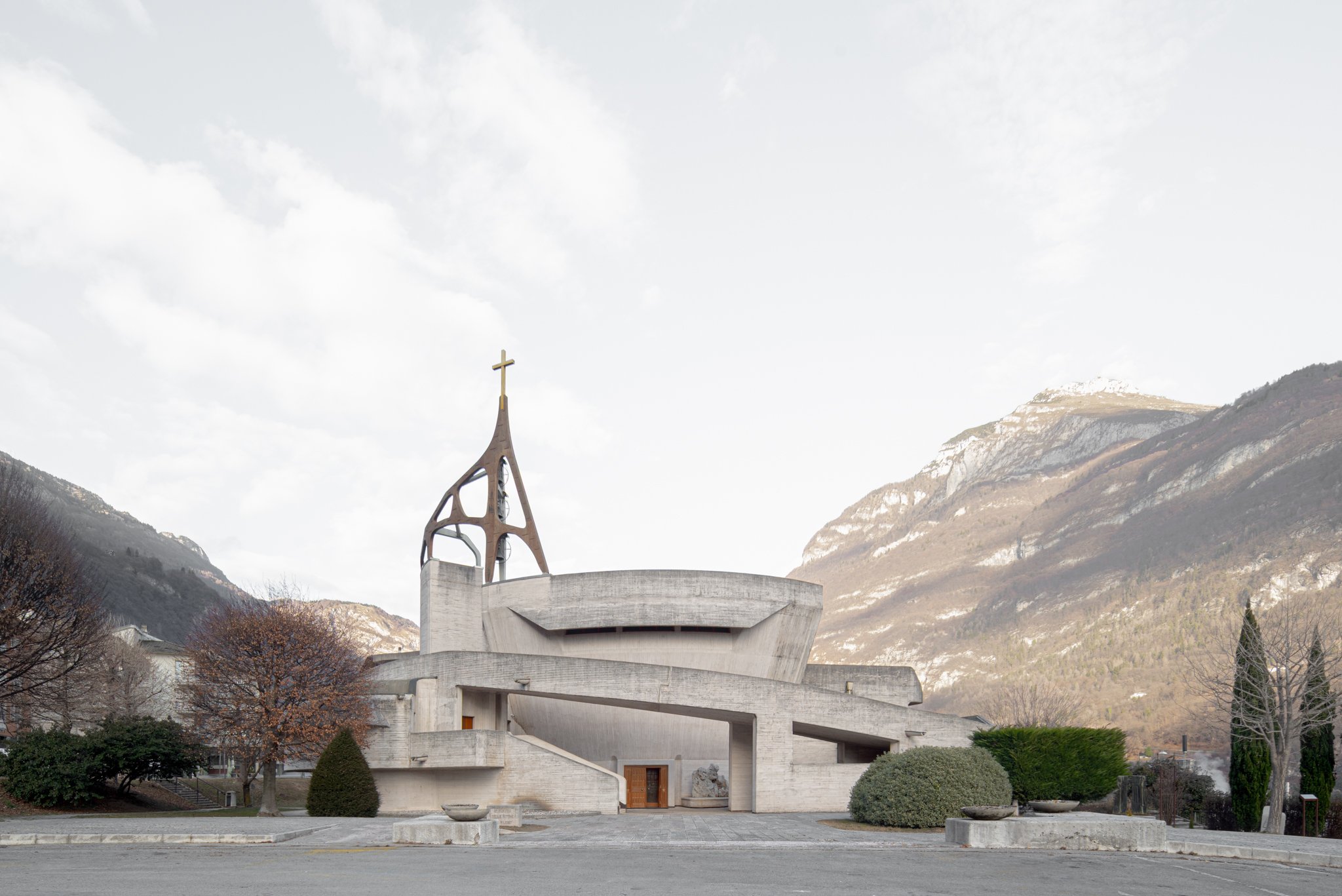 Chiesa moderna con architettura in cemento e una torre con una croce, circondata da montagne e alberi