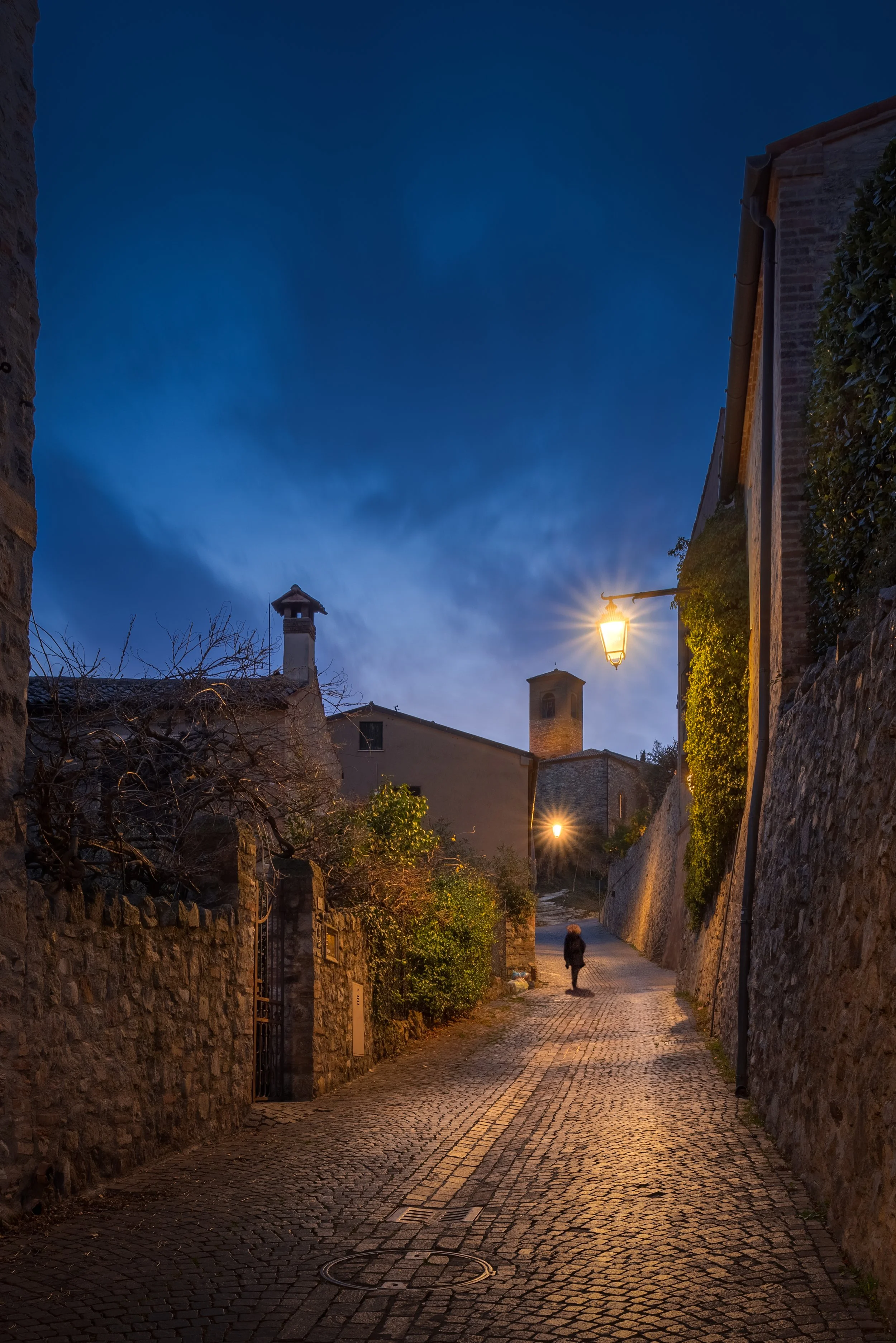 Veduta di una strada di ciottoli in una zona storica di notte, con luci di lampioni e una persona che cammina sul marciapiede, case con muri di pietra e tetti di tegole, cielo scuro con nuvole.