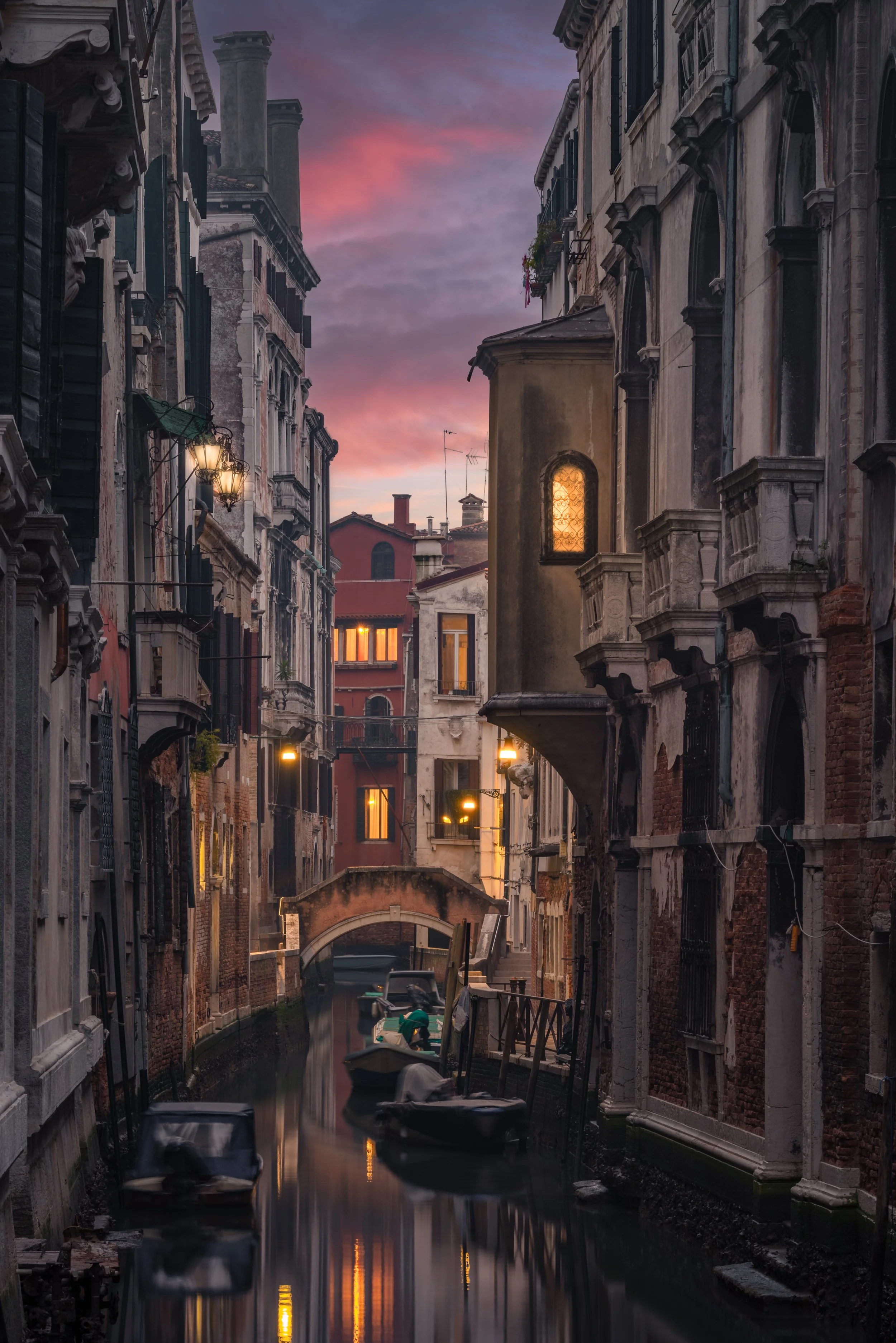Veduta di un canale di Venezia con barche ormeggiate ai lati, edifici antichi con finestre illuminate e un piccolo ponte pedonale, al tramonto con cielo colorato di rosa e viola.
