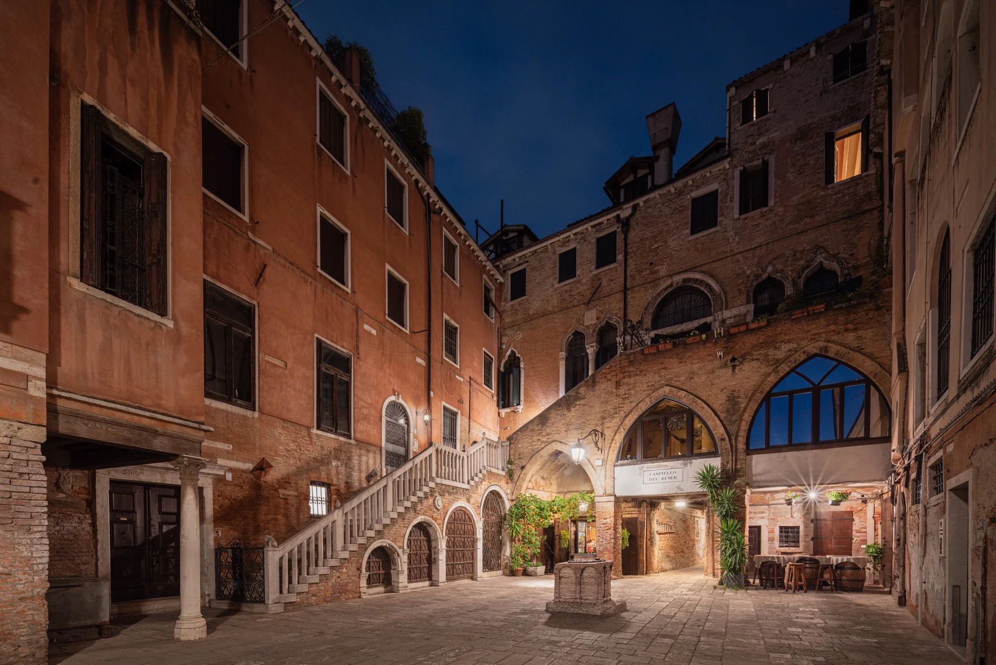 Vicolo di Venezia di sera con edifici storici in mattoni, scale in pietra, piante e tavoli all'aperto.
