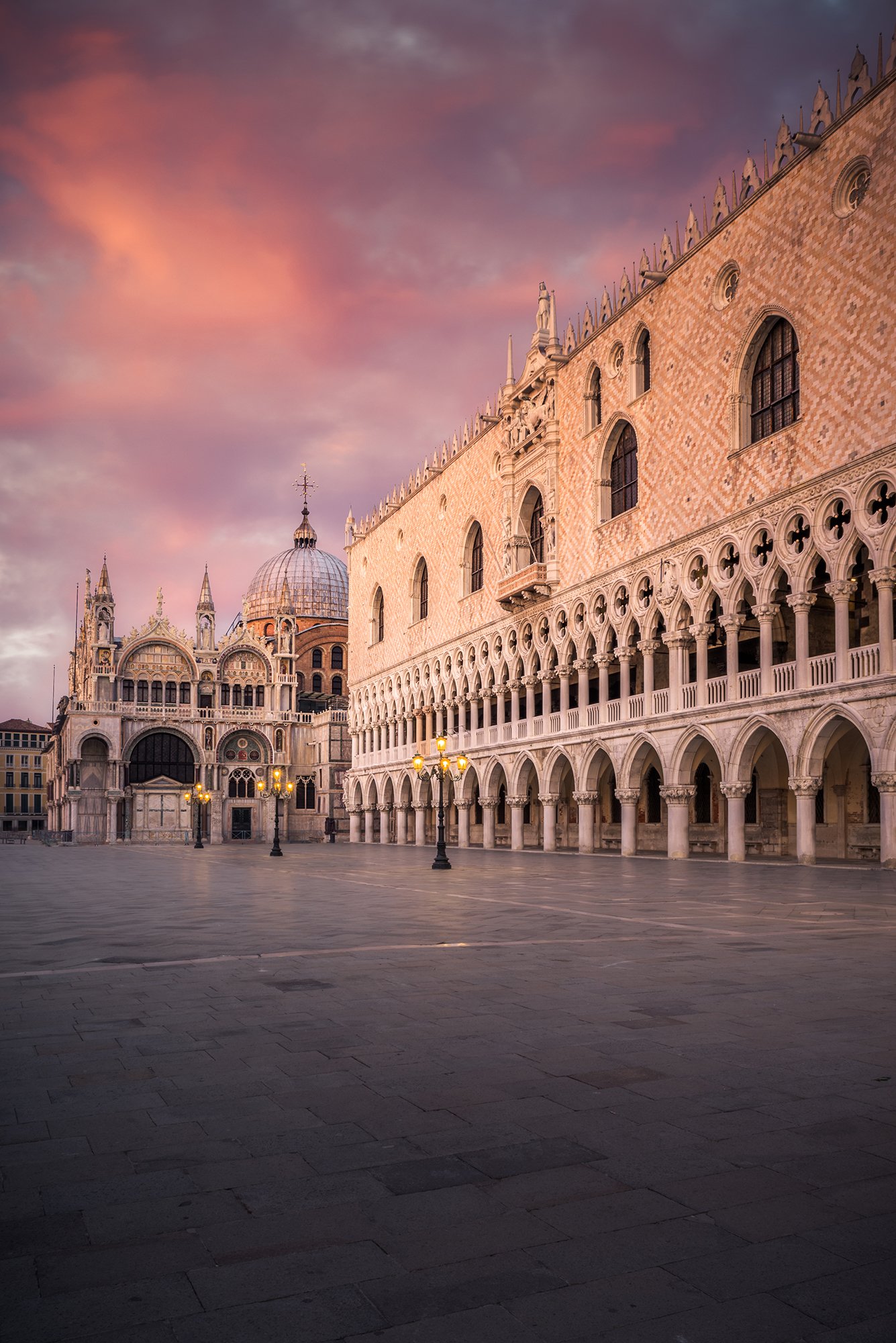 Veduta della piazza di San Marco a Venezia al tramonto, con il Duomo di San Marco e il Palazzo Ducale visibili sullo sfondo, cielo colorato di rosa e viola, lampioni accesi.