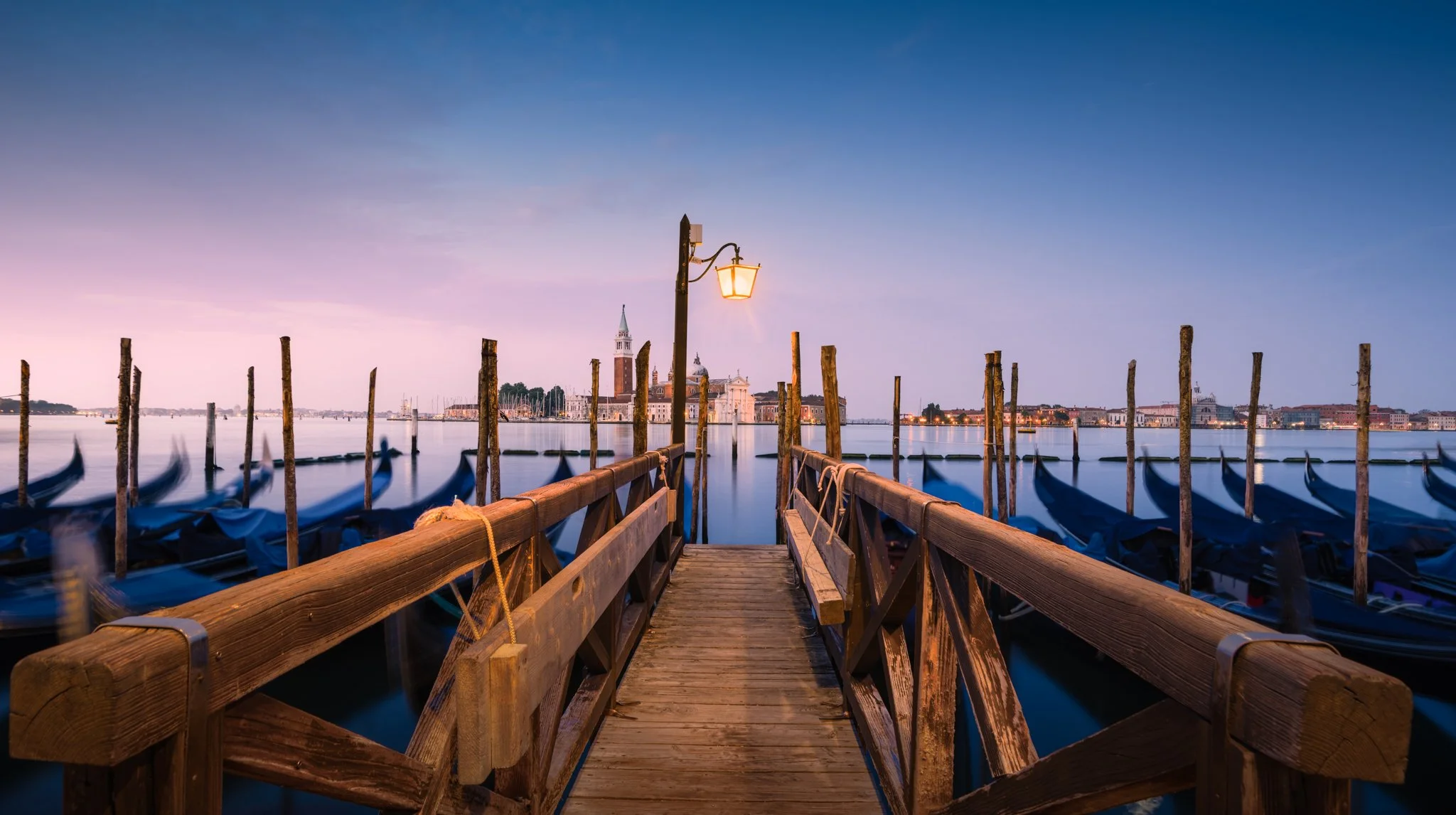 Molo di legno con lampione acceso che guarda verso Venezia con gondole ormeggiate e campanile di San Marco sullo sfondo al tramonto.