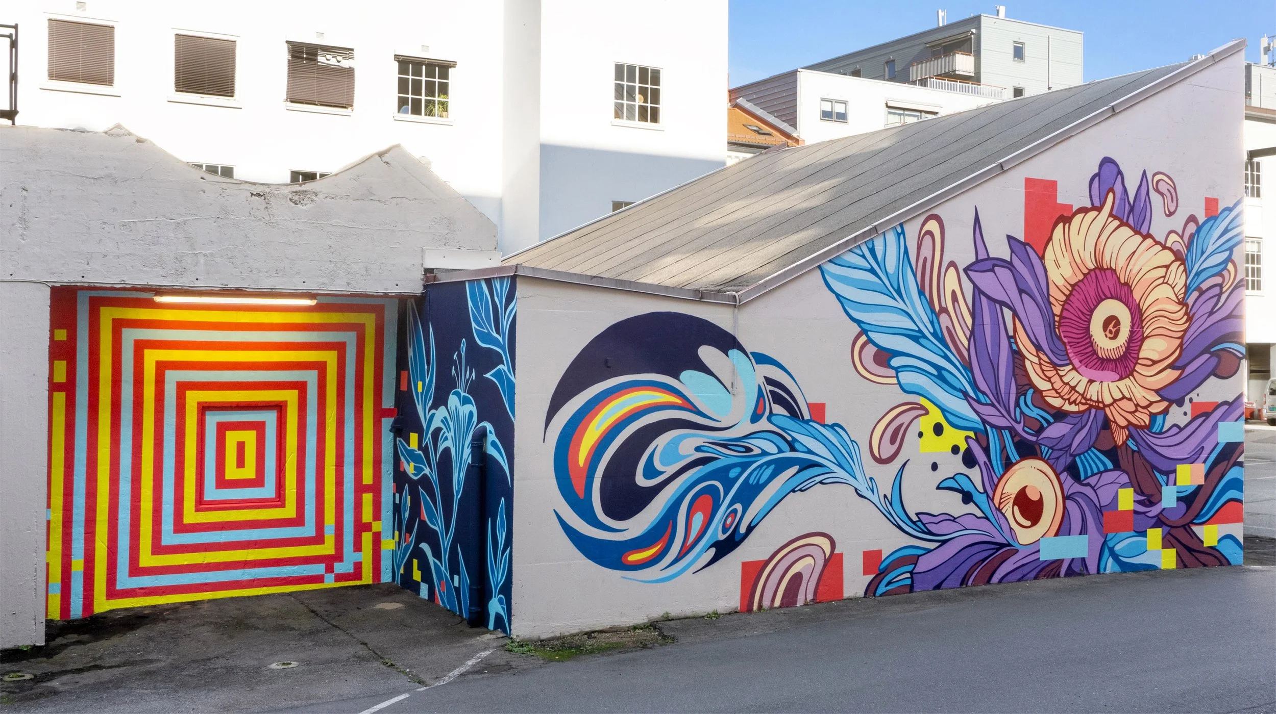 Colorful mural on a building wall, featuring a whale with water and rainbow patterns, surrounded by abstract and floral designs, including a large flower with purple leaves.
