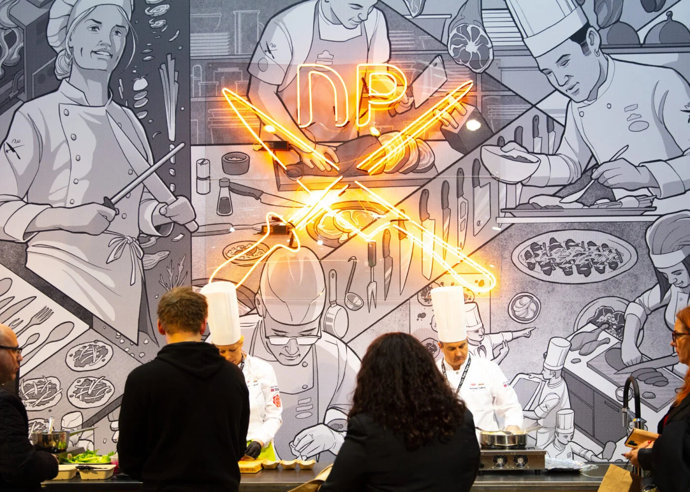 A culinary demonstration with professional chefs in front of a large wall mural depicting a grayscale cartoon kitchen scene. The mural features chefs preparing various dishes, with a neon sign that reads 'NDP' and stylized neon swords crossing throug