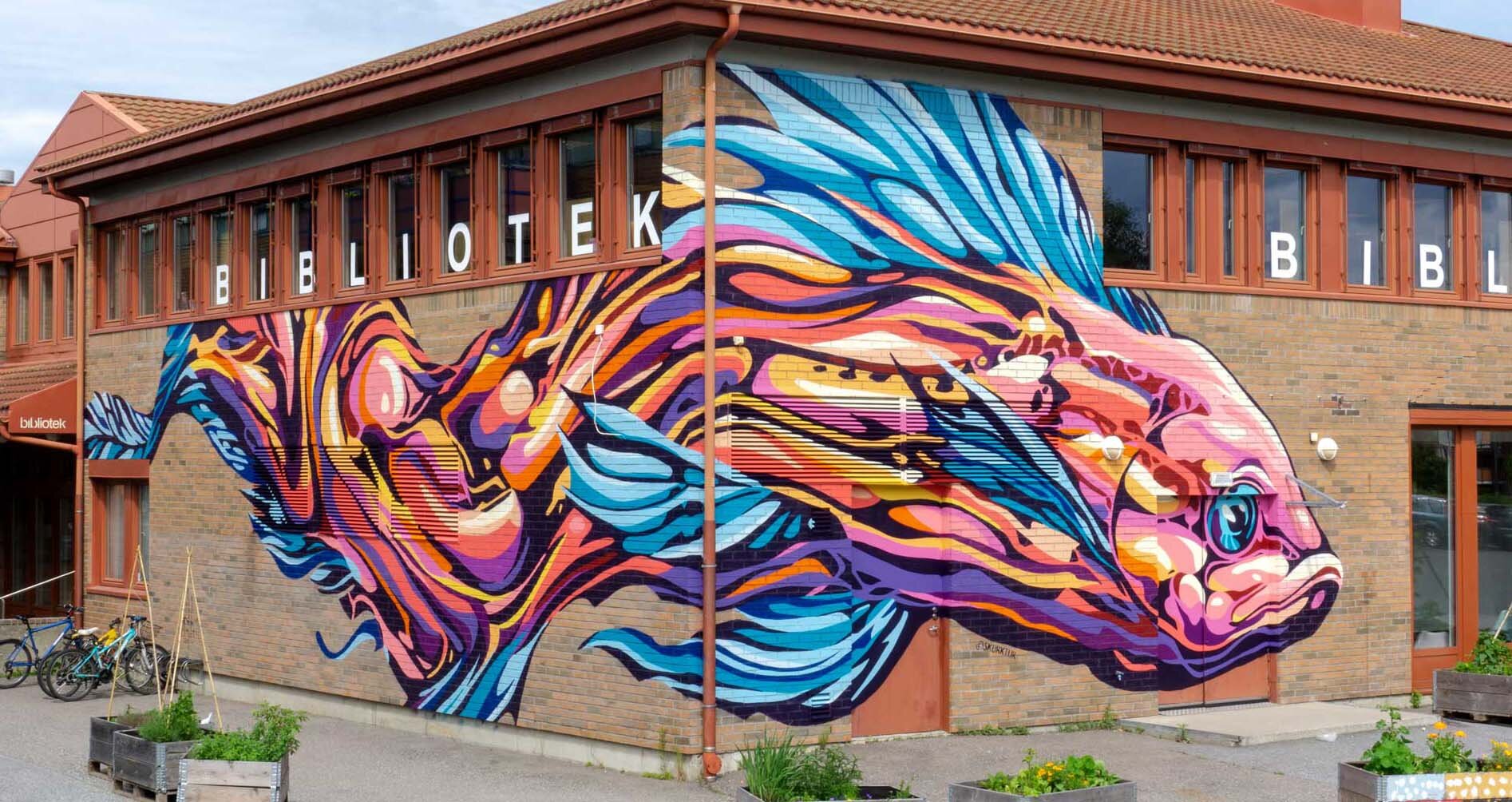 Colorful mural of a fish painted on the brick wall of a library building named 'BiblioTek' with bicycles and planters in front.