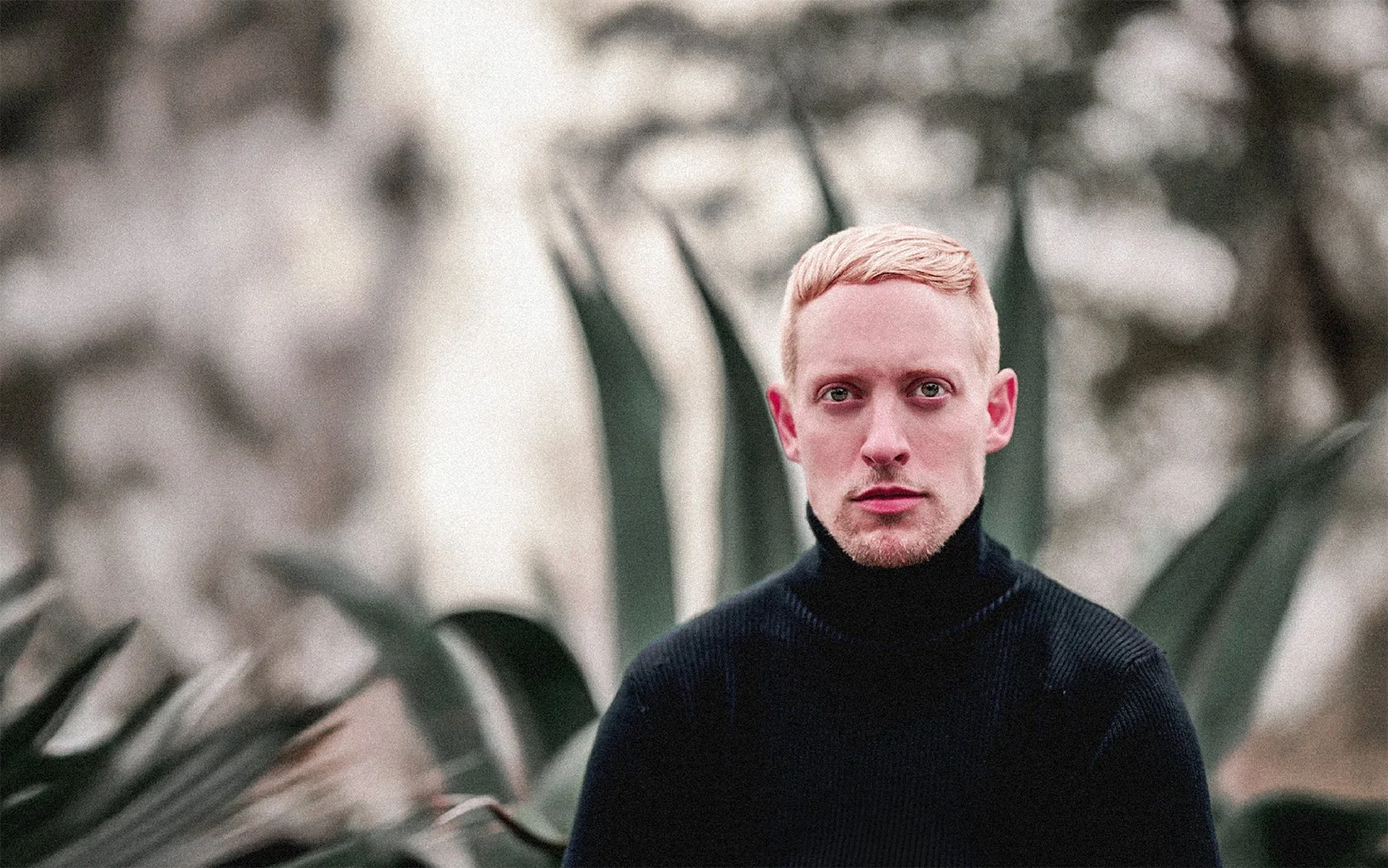 Michael Nordstrand, perfumer,  with short blond hair, wearing a black turtleneck sweater, standing in front of a blurred background of trees and large leaves, looking directly at the camera. – D:SOL